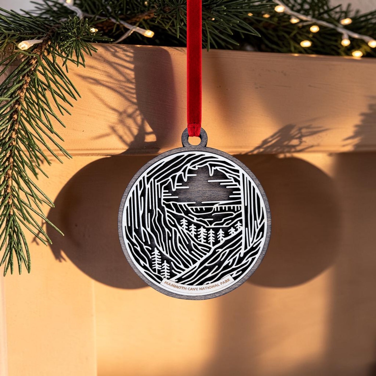 The Mammoth Cave National Park Line Art Christmas Ornament by Mirabilia Boutique, featuring an oval wooden design with a cave and tree motif, hangs from a red ribbon on a mantle adorned with pine branches and string lights.