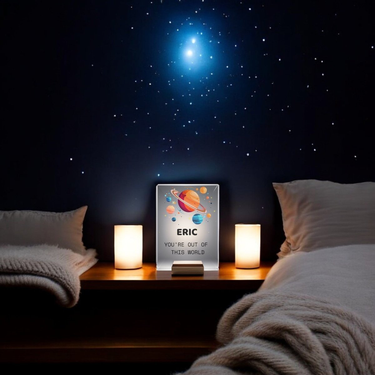 A cozy, space-themed bedroom with two beds, soft blankets, a wooden nightstand topped with two lamps and the Mirabilia Boutique Personalized Space Night Light for Kids, while glowing stars shine on the dark wall and a sign says "ERIC YOU'RE OUT OF THIS WORLD.
