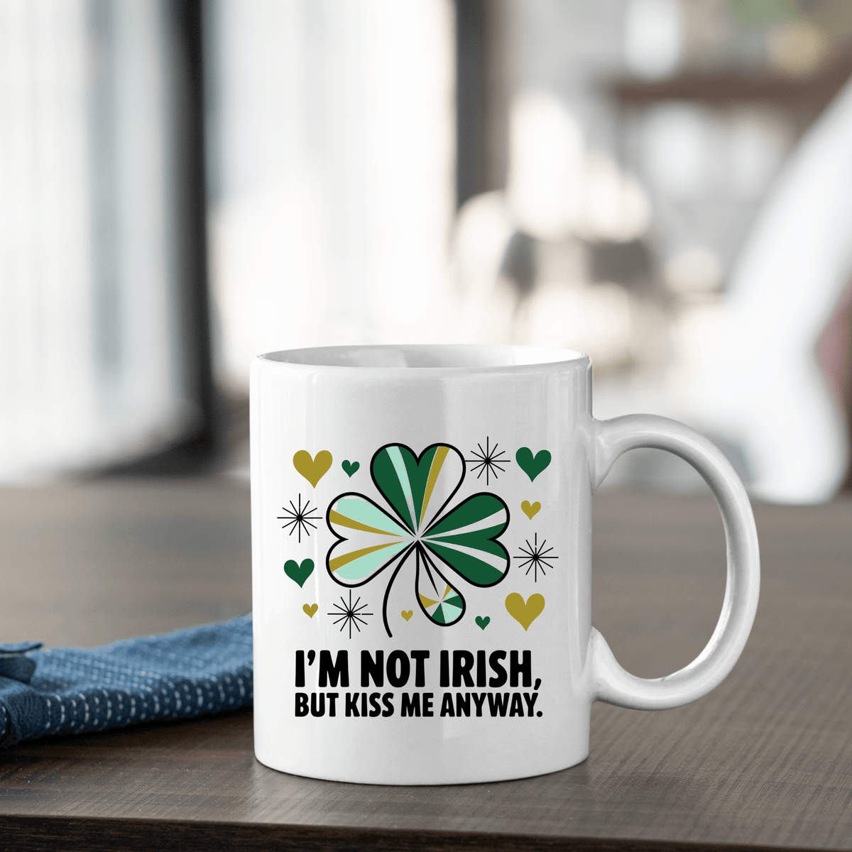 The Kiss Me Anyway Mug – St. Patrick’s Day Charm by Mirabilia Boutique sits on a wooden table, featuring a green shamrock, yellow and green hearts, and “I’M NOT IRISH, BUT KISS ME ANYWAY.” A folded blue cloth rests nearby.