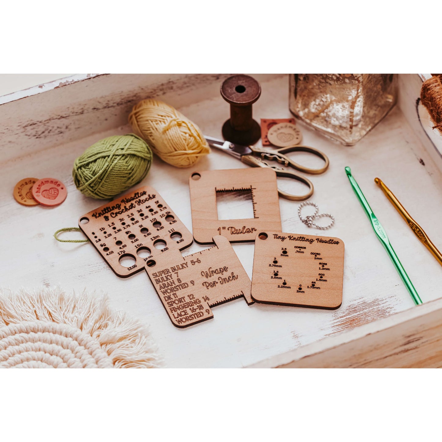 The Knitter’s Pocket Tool Set by Mirabilia Boutique features 4 mini tools with engraved measurements and patterns on a white tray. Accompanied by two yarn balls, crochet hooks, and a spool, it creates a cozy atmosphere perfect for travel knitting adventures.