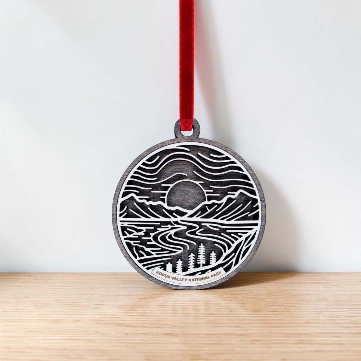 A round wooden ornament from Mirabilia Boutique hangs on a red ribbon, featuring line art of mountains, trees, a winding river, and a setting sun reminiscent of Alaska. Labeled “Kobuk Valley National Park,” this handcrafted Christmas ornament rests against a light wood background.