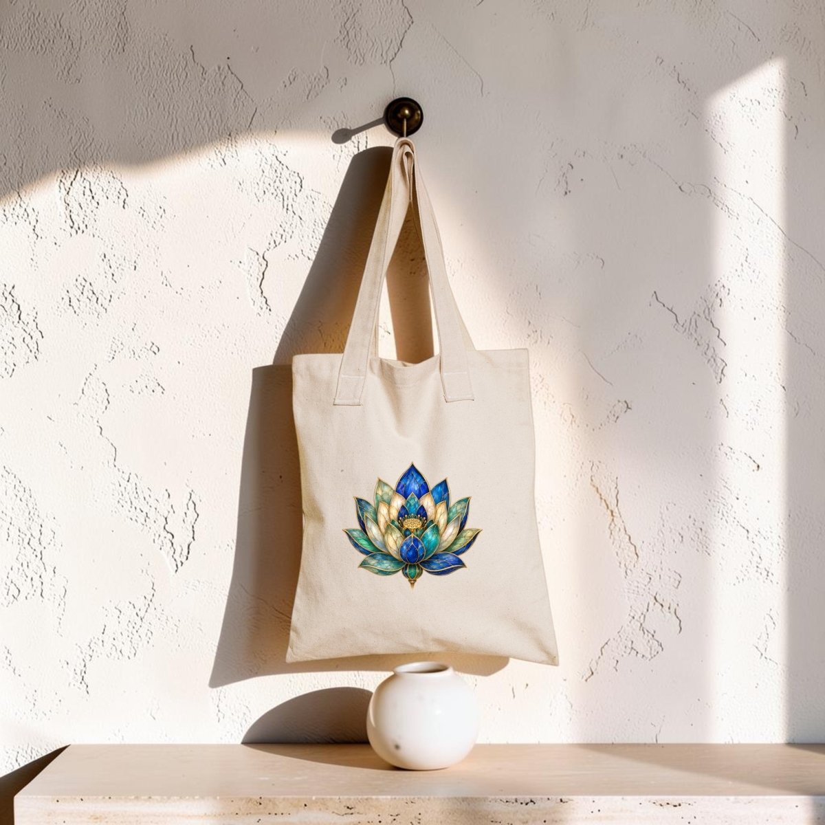 A Lapis Bloom Tote Bag | Egyptian Lotus Art from Mirabilia Boutique, featuring a colorful lotus flower design on beige fabric, hangs on a wall hook above a small white vase, with sunlight casting shadows across the textured wall.