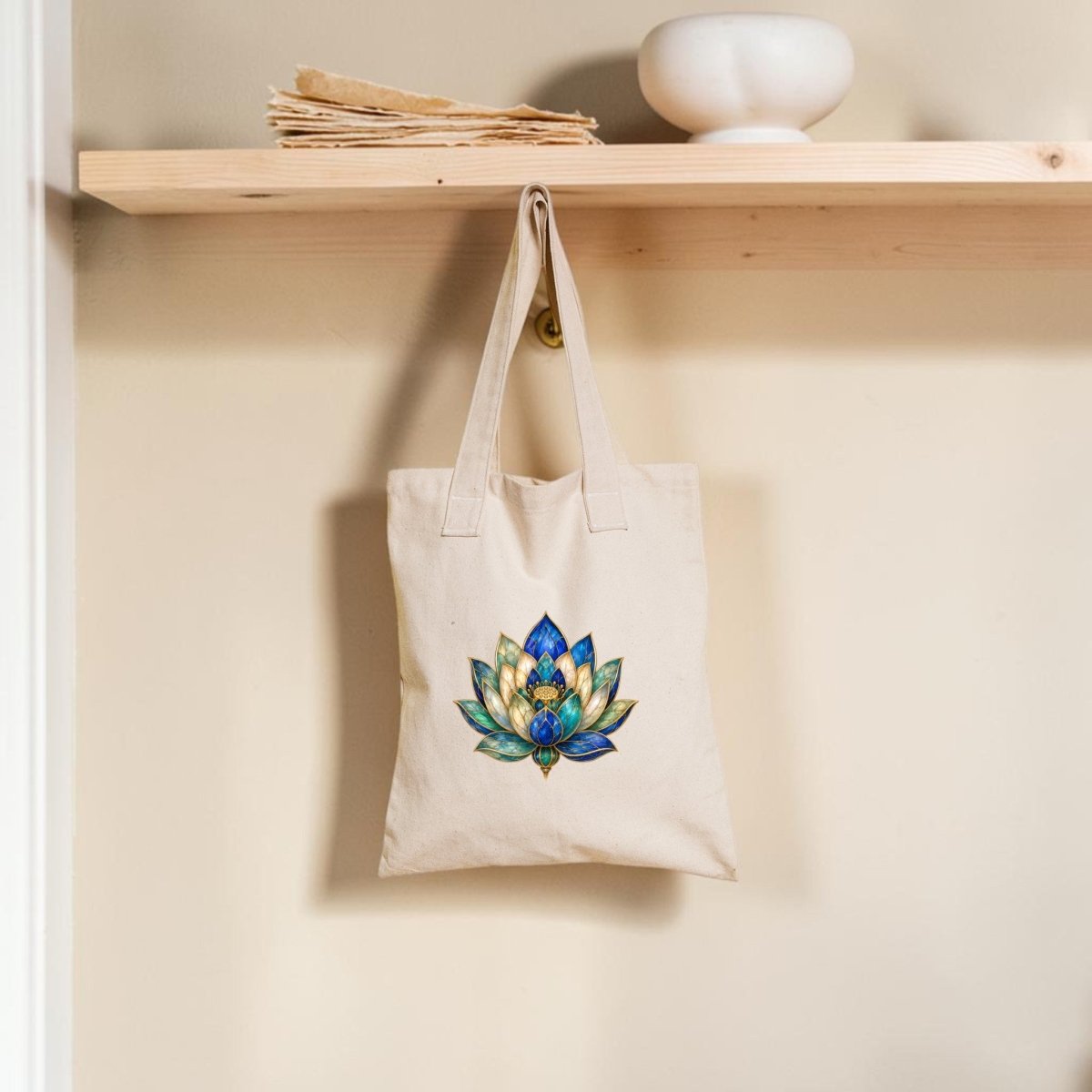 A Lapis Bloom Tote Bag | Egyptian Lotus Art by Mirabilia Boutique, featuring a colorful lotus flower design in beige, hangs from a wooden hook beneath a shelf with papers and a round white bowl against a cream wall.