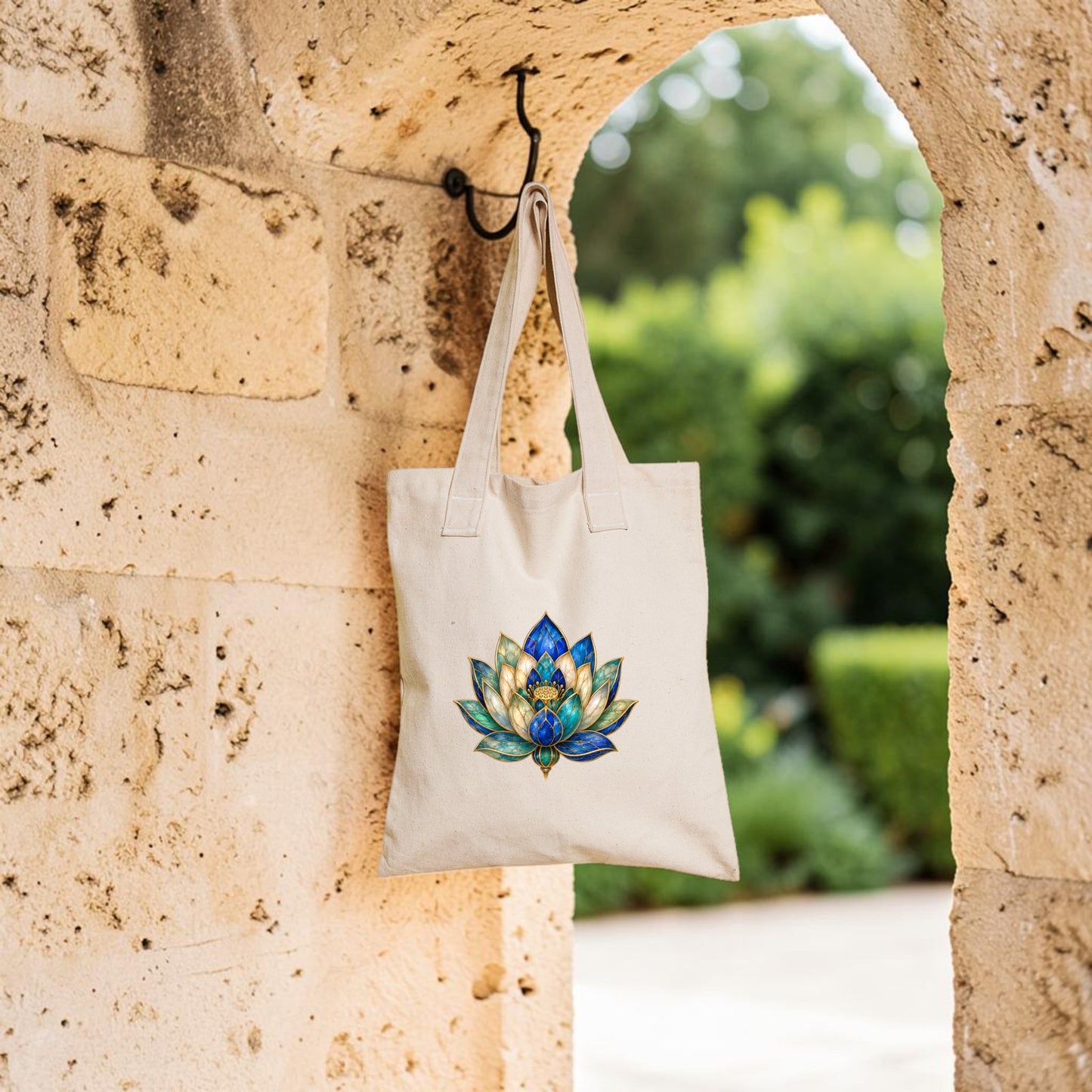 The Lapis Bloom Tote Bag | Egyptian Lotus Art by Mirabilia Boutique, featuring a vibrant lotus flower design on a beige background, hangs from a metal hook on a stone archway, with lush greenery softly blurred in the background.