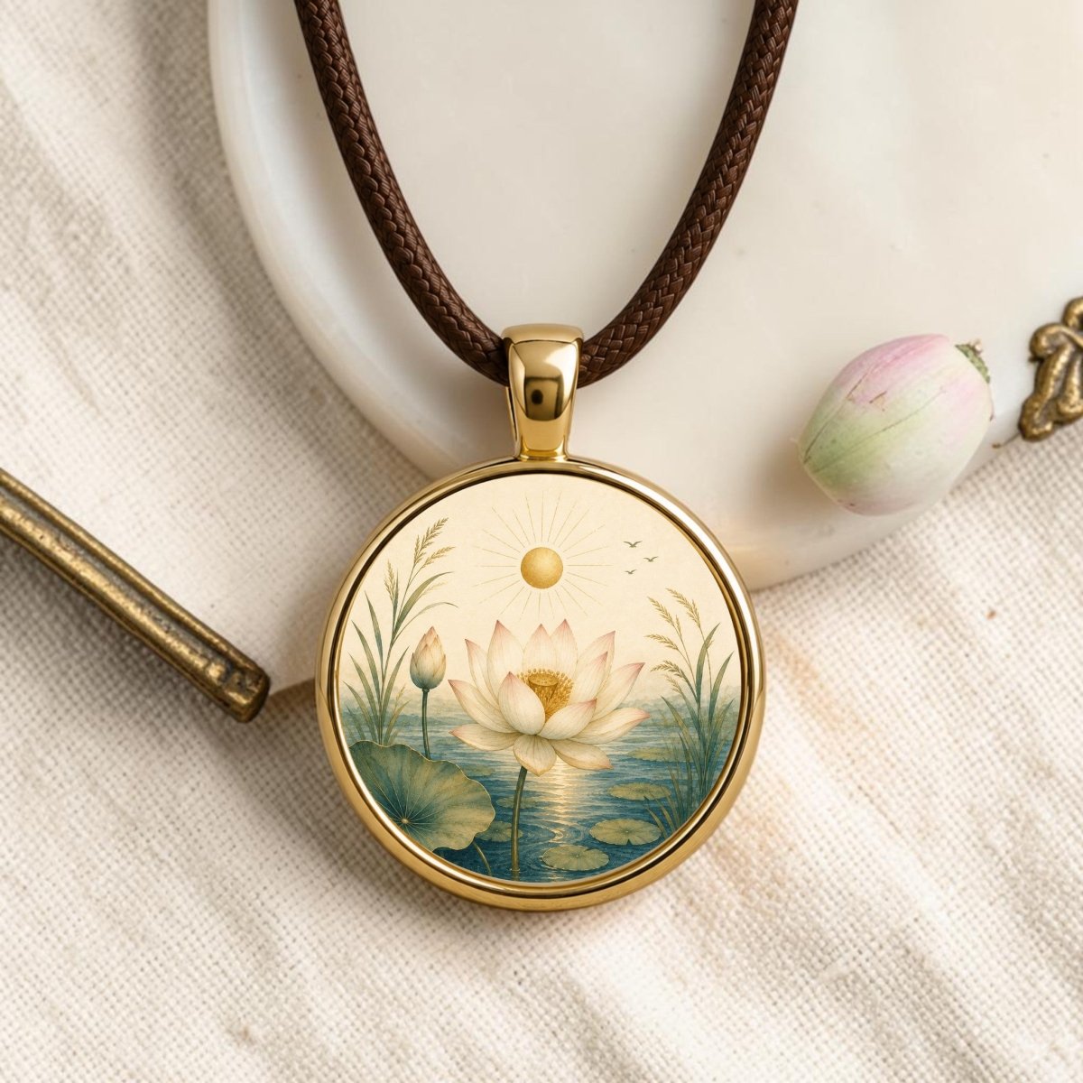 The Becoming | Story Pendant Necklace by Mirabilia Boutique is a gold lotus pendant on a brown cord, featuring an illustration of a white lotus flower and buds floating on water, surrounded by greenery, sunlight, and birds in the sky. This exquisite piece of botanical jewelry is displayed on textured cream fabric next to a pink flower bud.