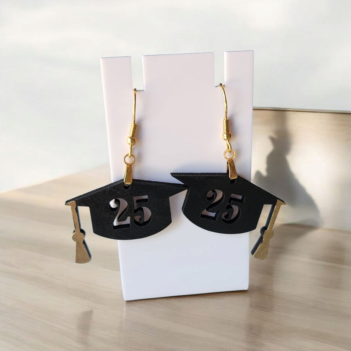 Mirabilia Boutique’s Graduation Cap Earrings Class of 2025 are laser-cut black cap earrings with gold tassels and “25”—a perfect grad jewelry gift for her or party accessory. Shown on a white stand atop a wooden surface.