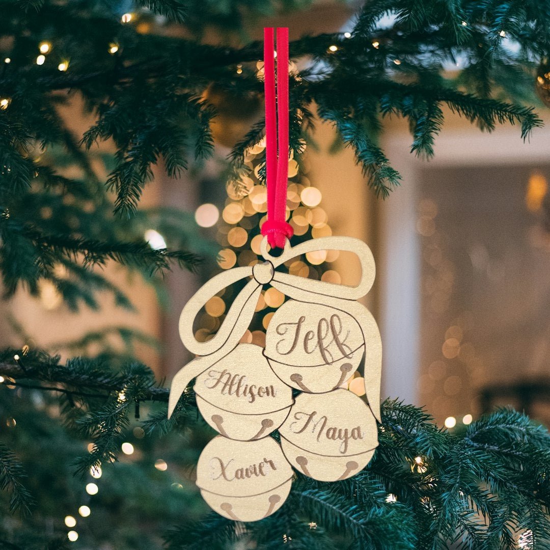 The Mirabilia Boutique Custom Family Christmas Ornament—a laser-cut wooden jingle bell with a bow—features the names Jeff, Allison, Maya, and Xaver, hanging on a tree amid twinkling holiday lights.