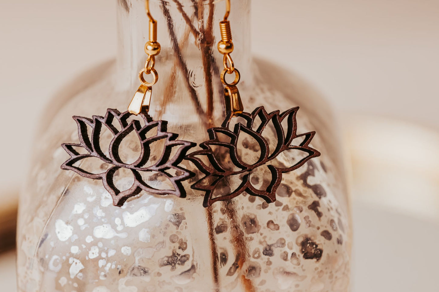 A close-up of Mirabilia Boutique’s Laser Cut Lotus Flower Wood Earrings—lightweight, boho-style pieces with gold hooks—hanging on a textured glass bottle, showcasing their delicate outlined design.