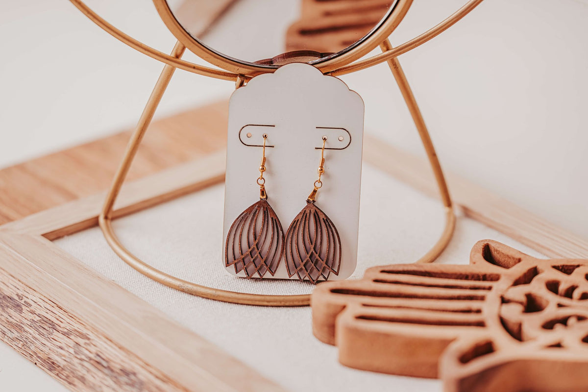 A pair of brown, leaf-shaped Laser Cut Tulip Earrings by Mirabilia Boutique with hypoallergenic hooks displayed on a white card atop a wooden tray, surrounded by decorative accents and a round mirror in the background.