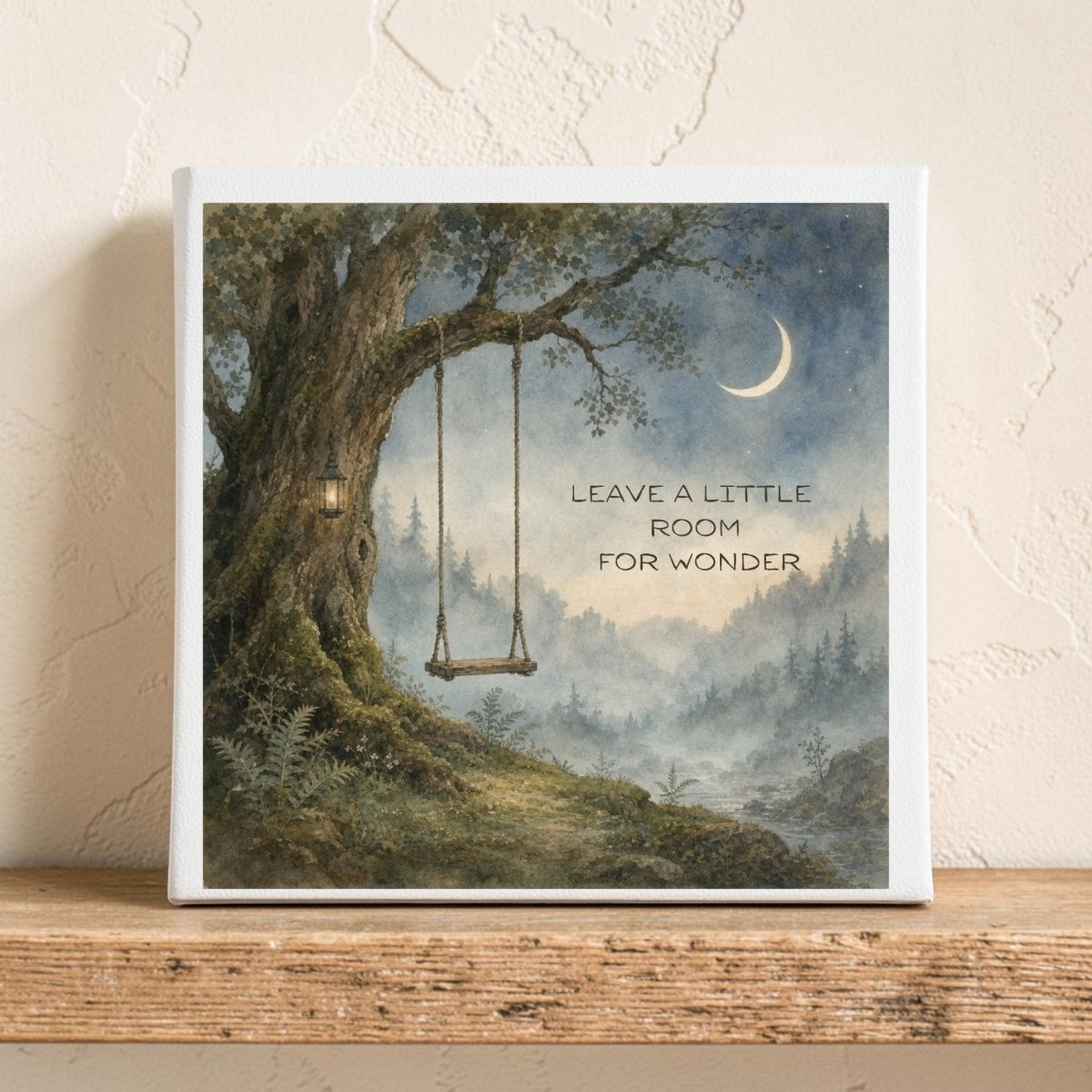 The Leave a Little Room for Wonder Canvas by Mirabilia Boutique features a watercolor landscape with misty mountains, a towering tree with a swing, and a crescent moon. The artwork is adorned with the inspiring words, "LEAVE A LITTLE ROOM FOR WONDER." Resting elegantly on a wooden shelf against a textured wall, this piece brings nature-inspired charm to any room.