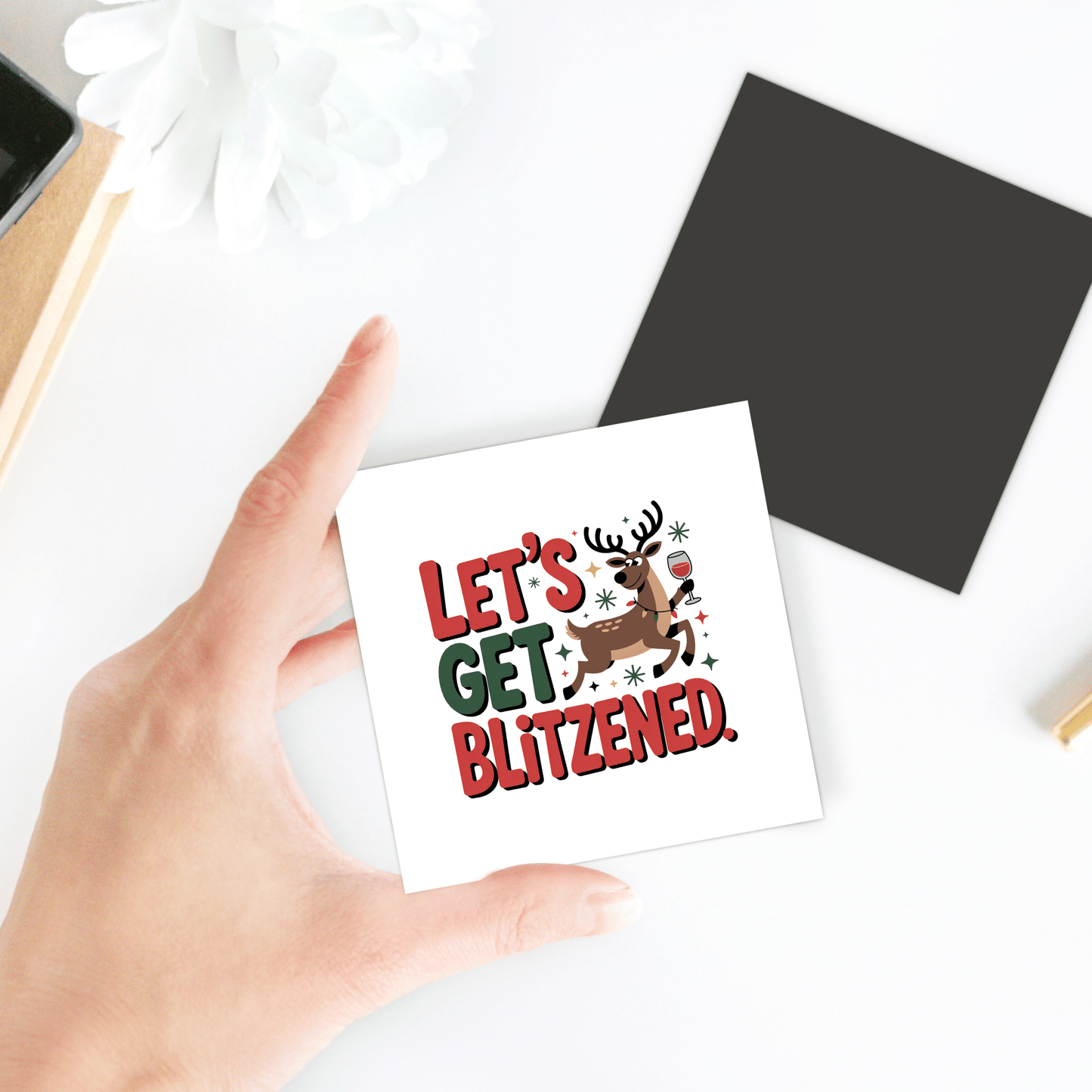 A hand holds the Let's Get Blitzened Magnet by Mirabilia Boutique, featuring a festive reindeer with a drink and "LET'S GET BLITZENED" in red and green—perfect funny Christmas fridge decor or gift for wine lovers.