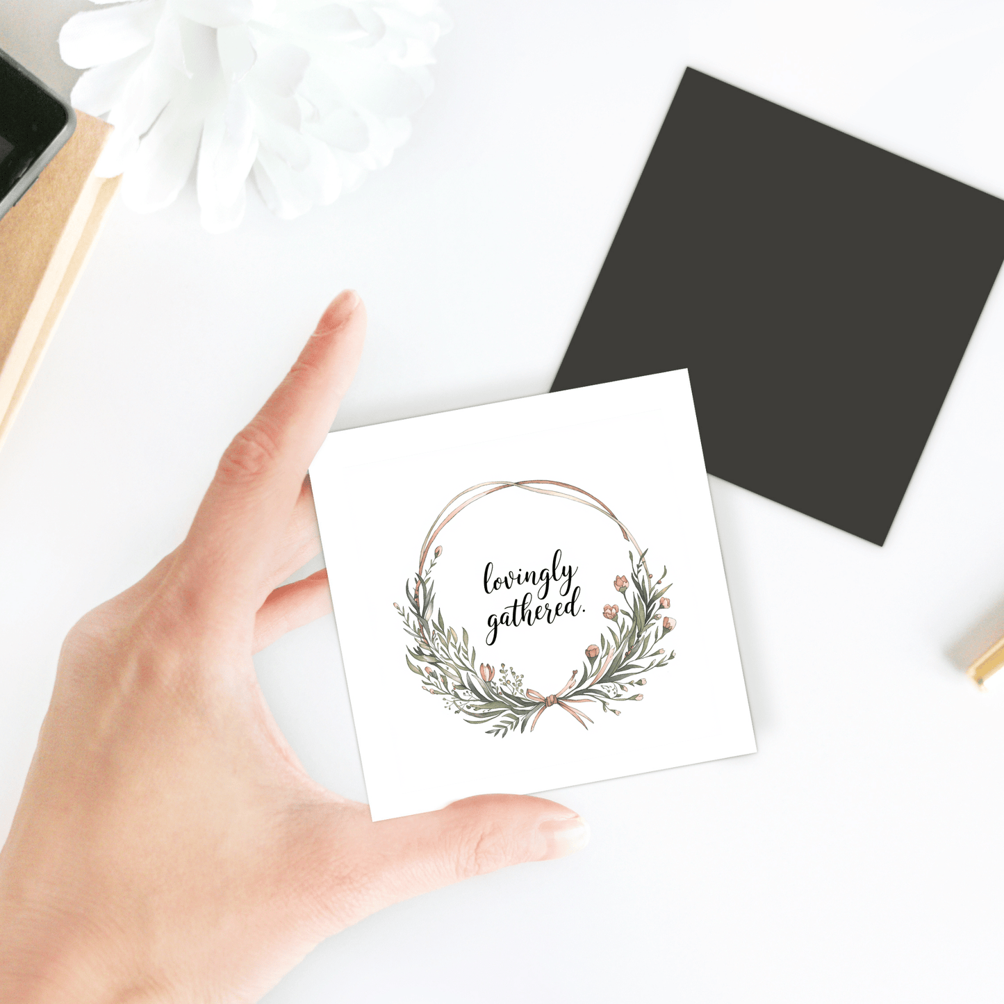 A hand holds a white card featuring a wreath and “lovingly gathered” beside a Mirabilia Boutique Lovingly Gathered Magnet – Floral Wreath Quote for Fridge, a black envelope, and a white flower on a white surface.