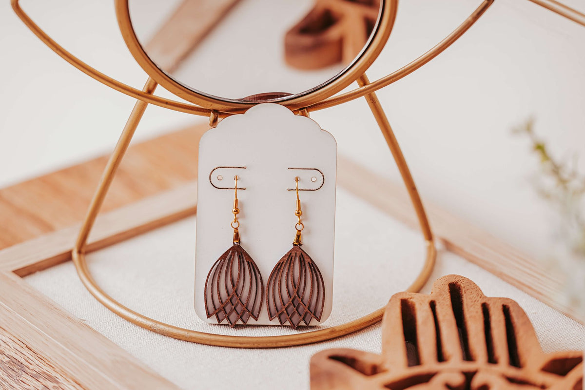 Mirabilia Boutique’s Laser Cut Tulip Earrings—lightweight, brown and gold boho dangles—are shown on a white card in a gold jewelry stand, set against wood accents and a soft, neutral background. Hypoallergenic hooks included.