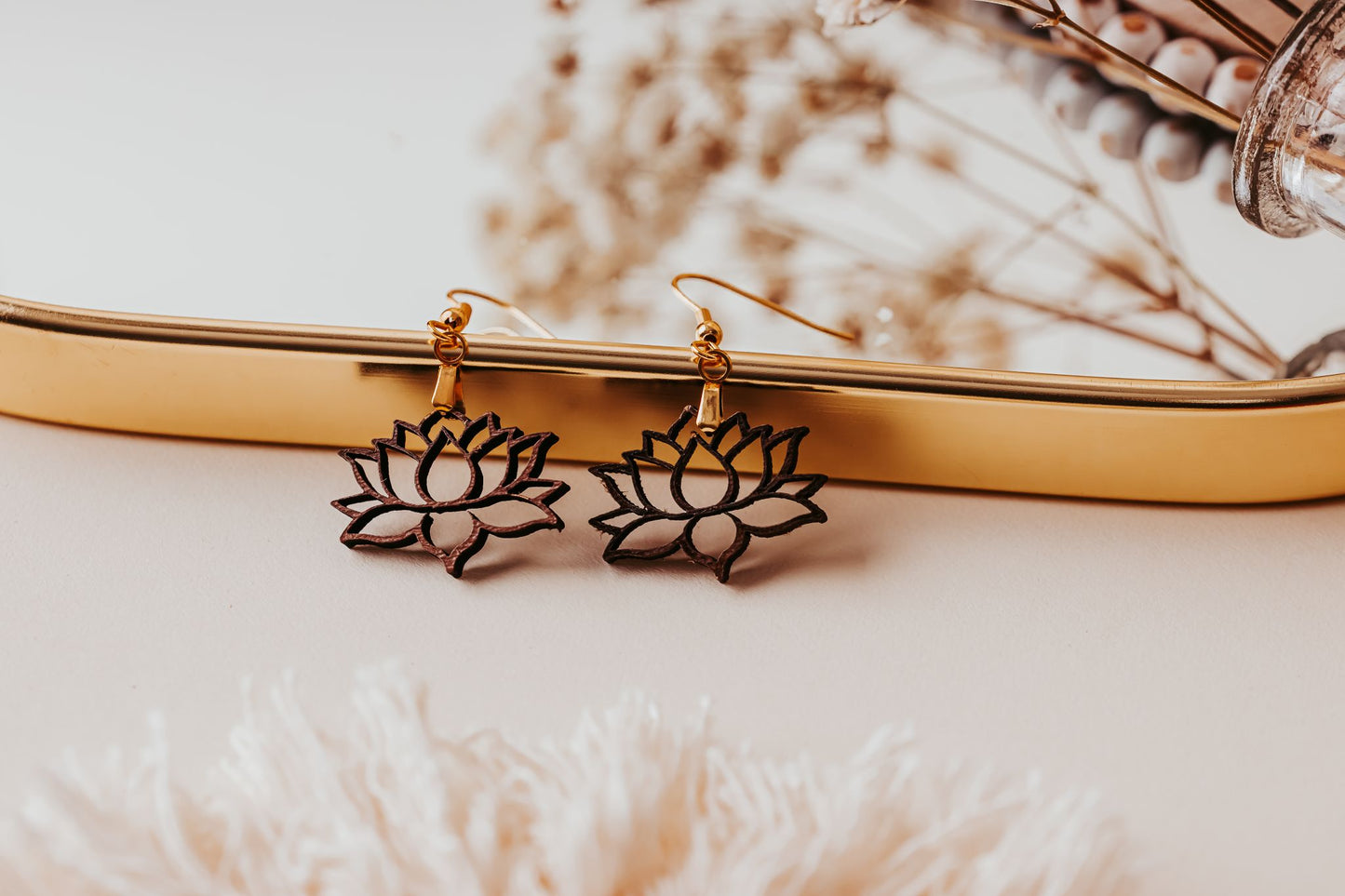 Mirabilia Boutique’s Laser Cut Lotus Flower Wood Earrings with gold hooks rest on a gold tray, surrounded by dried flowers and soft fabric, adding a lightweight and delicate boho touch.