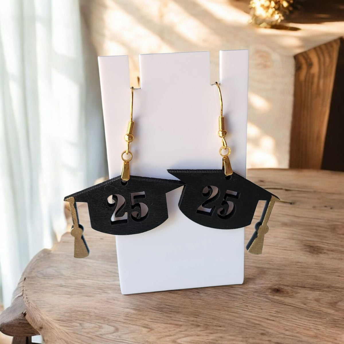 Mirabilia Boutique’s Graduation Cap Earrings Class of 2025—laser cut, with gold hooks and “25” detail—make a perfect grad gift for her. Shown displayed on a white stand atop a wooden table, softly lit by natural light.