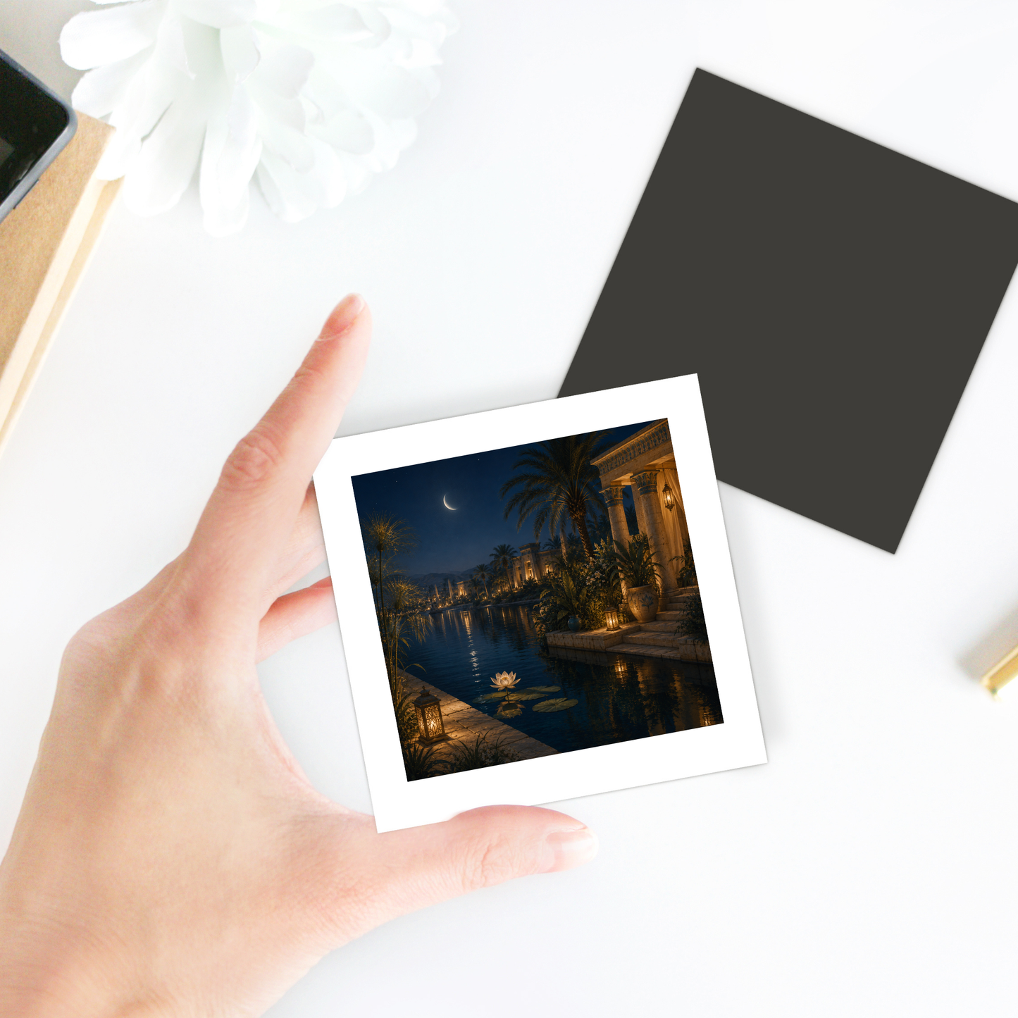 A hand holds a photo showing a night scene with a crescent moon, palm trees, a lit pool, and a villa. In the background sits the Lotus After Dark Magnet from Mirabilia Boutique—an Egyptian magnet shaped like an elegant lotus flower—alongside a white flower, black square card, and other desk items.