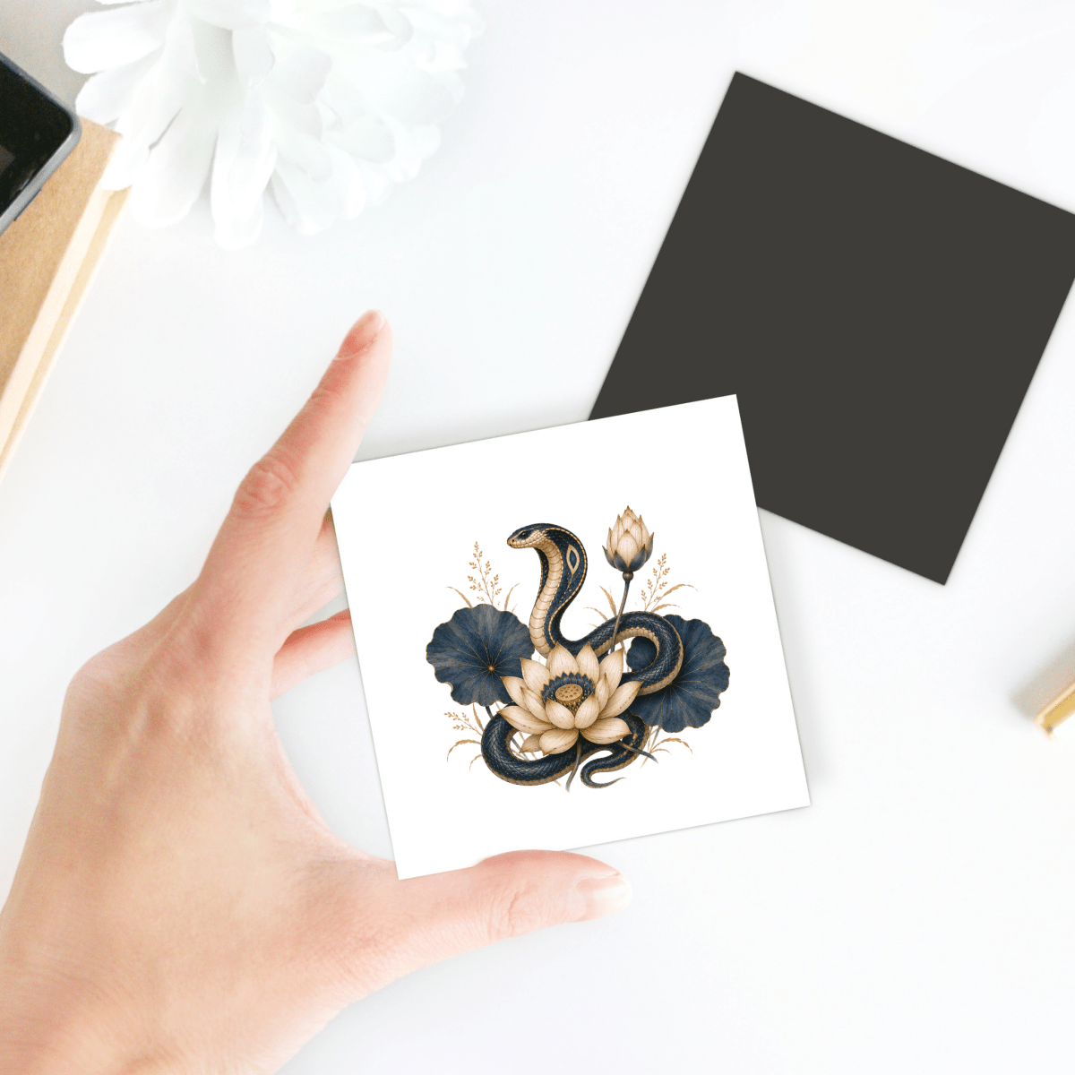 A hand holds the Lotus Guardian Cobra Magnet by Mirabilia Boutique—a square magnet featuring a snake coiled around cream and dark blue flowers inspired by Lapis & Lotus Egyptian décor. In the background, a flower and a black square object rest on the white desk.