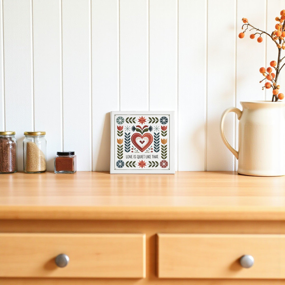 The Love Is Quiet Like That Canvas by Mirabilia Boutique, featuring folk art and a heartwarming quote, adds a charming and cozy touch to home decor on a white paneled wall.