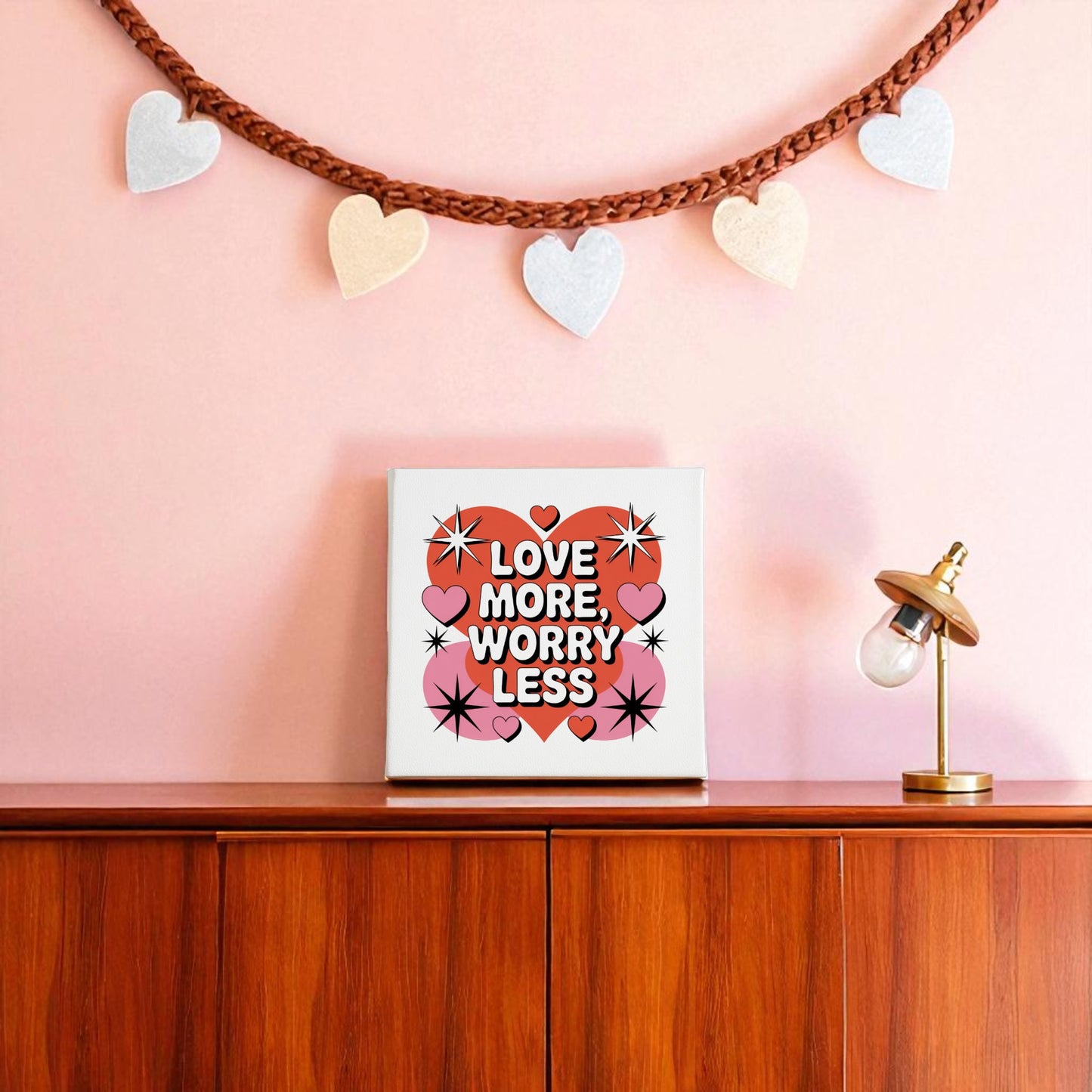 The Love More Worry Less Canvas by Mirabilia Boutique, an 8x8 midcentury wall art piece with a pink and red quote, sits on a wooden cabinet against a pink wall decorated with a heart garland, lamp, and retro accents.