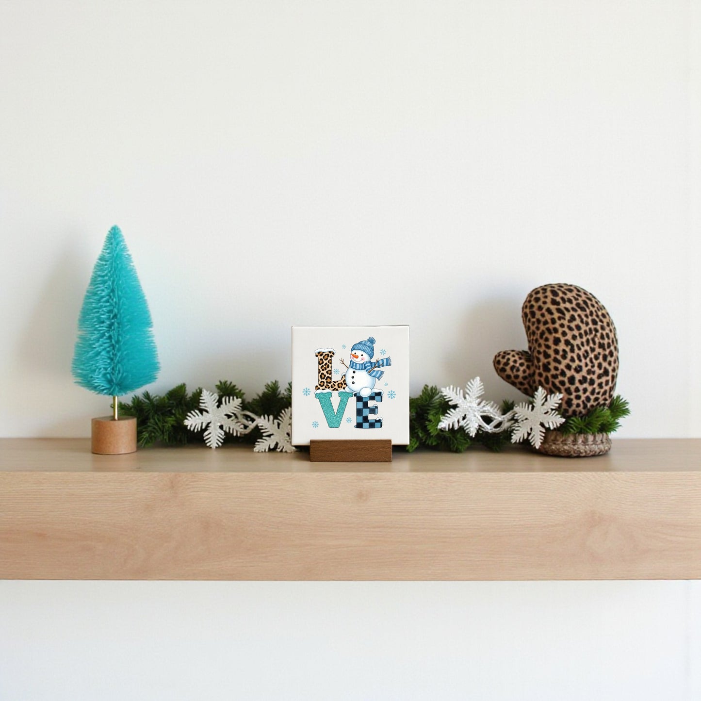 A minimalist wooden shelf displays winter decor: a blue bottle-brush tree, a Mirabilia Boutique Mini LOVE Snowman Sign, white faux pine garland, and a brown leopard-print mitten against a plain white wall.