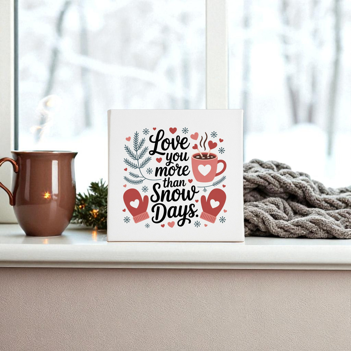 A ceramic mug and a knitted gray blanket sit on a windowsill beside Mirabilia Boutique’s "Love You More Than Snow Days" print—a charming winter wall art gift—while snowy trees outside set the perfect festive scene.