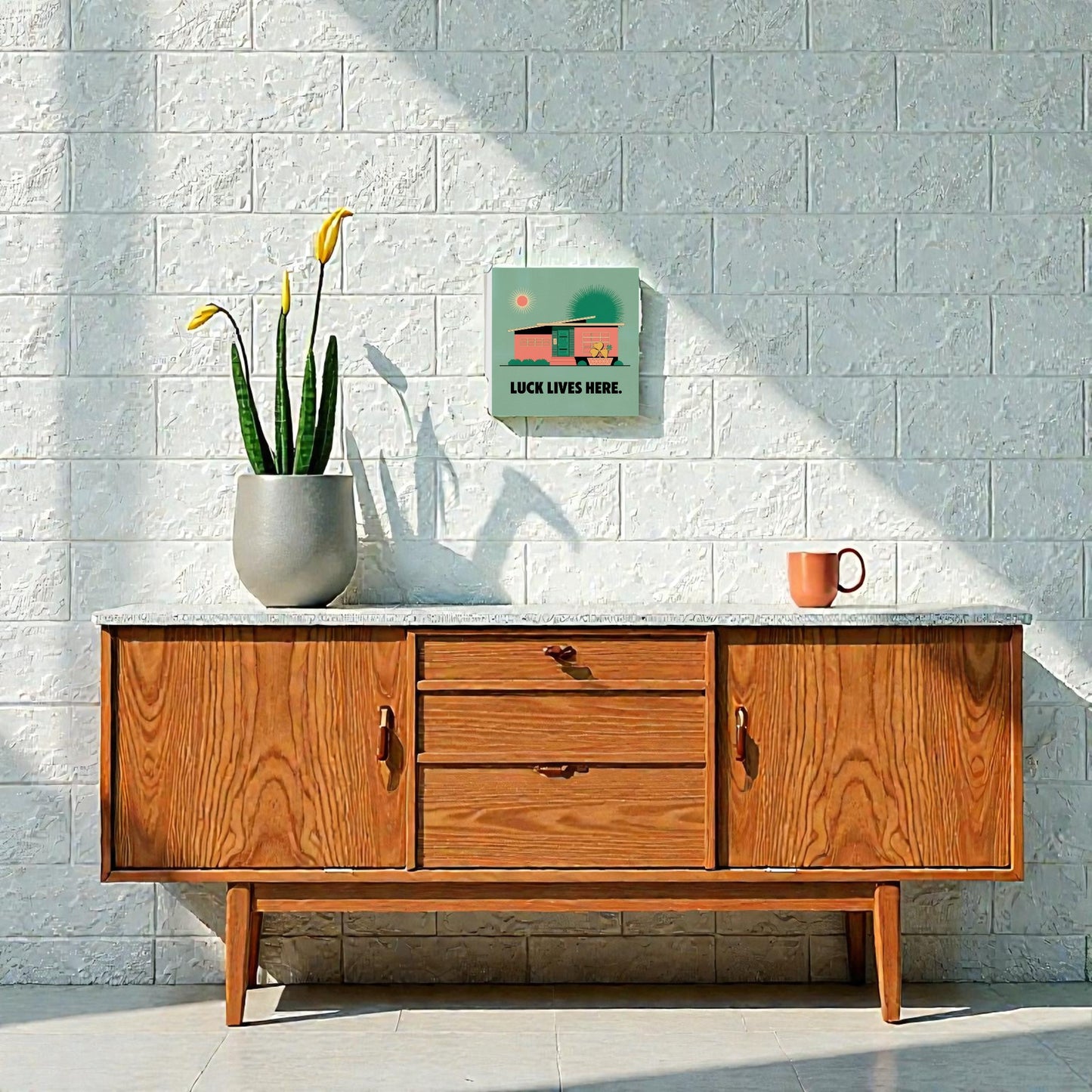 A wooden sideboard stands against a white brick wall, topped with a gray pot of yellow flowers and a pink mug. Above hangs the Mirabilia Boutique Luck Lives Here Canvas Sign – Midcentury Home Art, featuring green and orange accents.