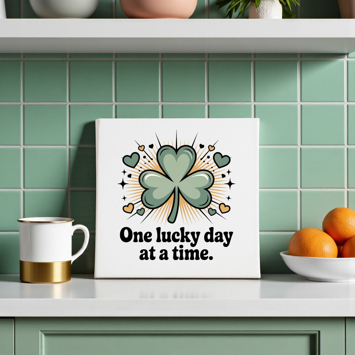 A kitchen with green tiled walls displays the Mirabilia Boutique's “One Lucky Day at a Time Canvas,” decorated with a shamrock, hearts, and sparkles. A mug and a bowl of oranges sit nearby on the counter.