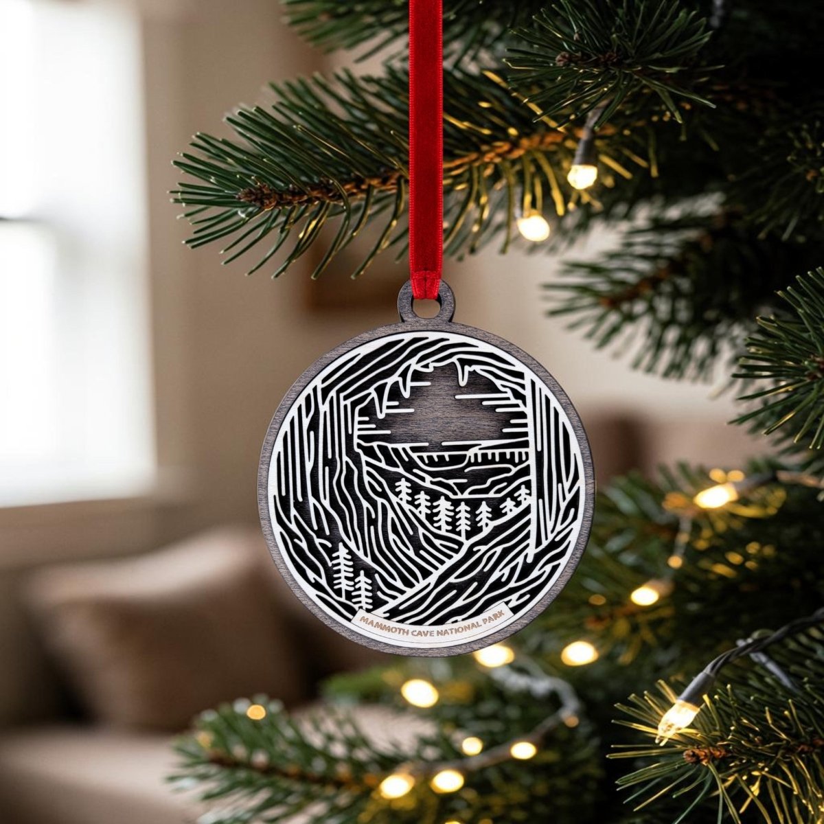 The Mammoth Cave National Park Line Art Christmas Ornament by Mirabilia Boutique is a handcrafted black and white wooden keepsake featuring a cave and forest design. It hangs from a red ribbon on a lit Christmas tree branch, set against a softly blurred indoor background.