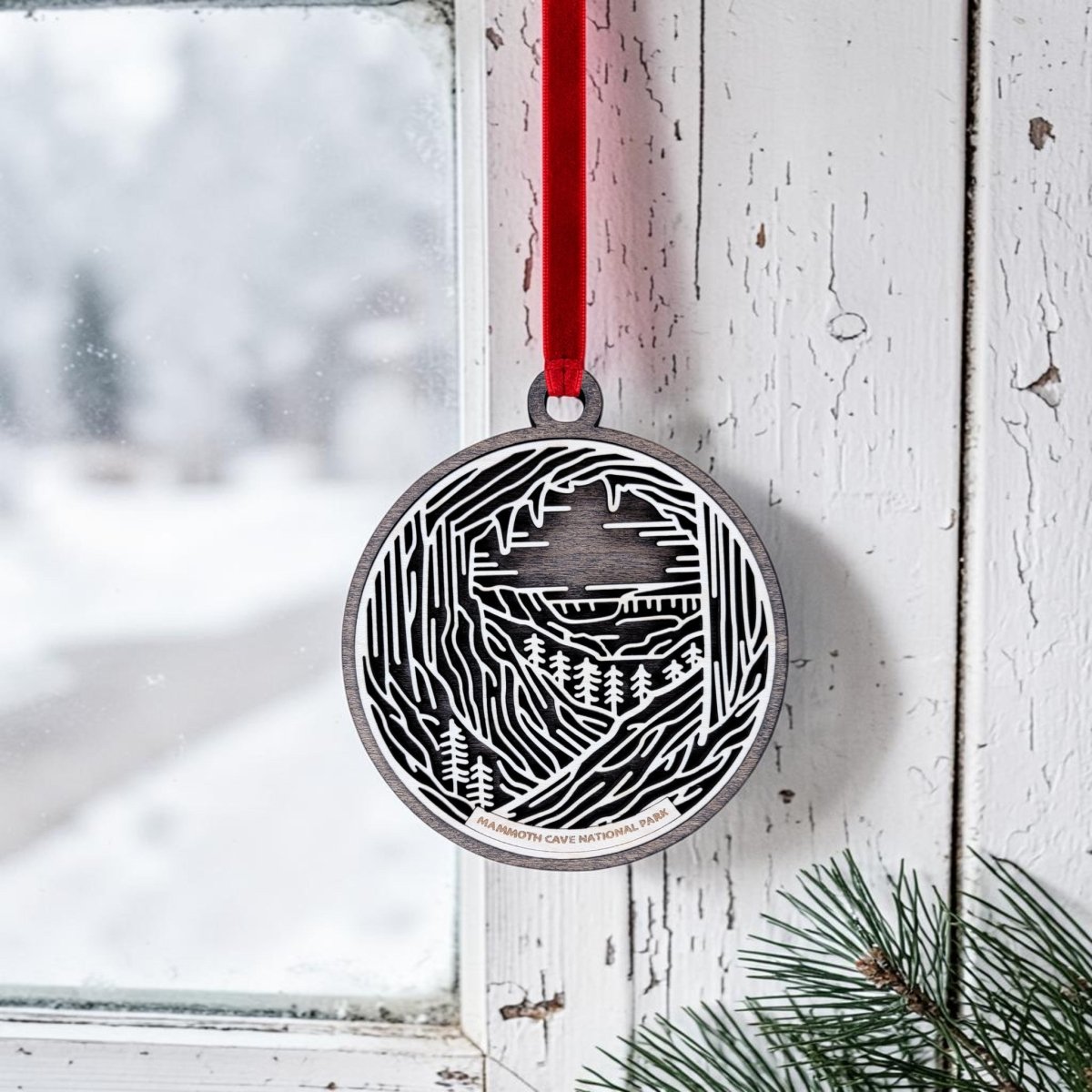 Celebrate the serene beauty of Mammoth Cave National Park with the Mammoth Cave National Park Line Art Christmas Ornament by Mirabilia Boutique. This handcrafted metal ornament, adorned with an elegant mountain and cave design, hangs from a red ribbon on a window frame. Snowy scenery is visible outside, while pine branches rest on the windowsill, capturing the peaceful spirit of the park during wintertime.