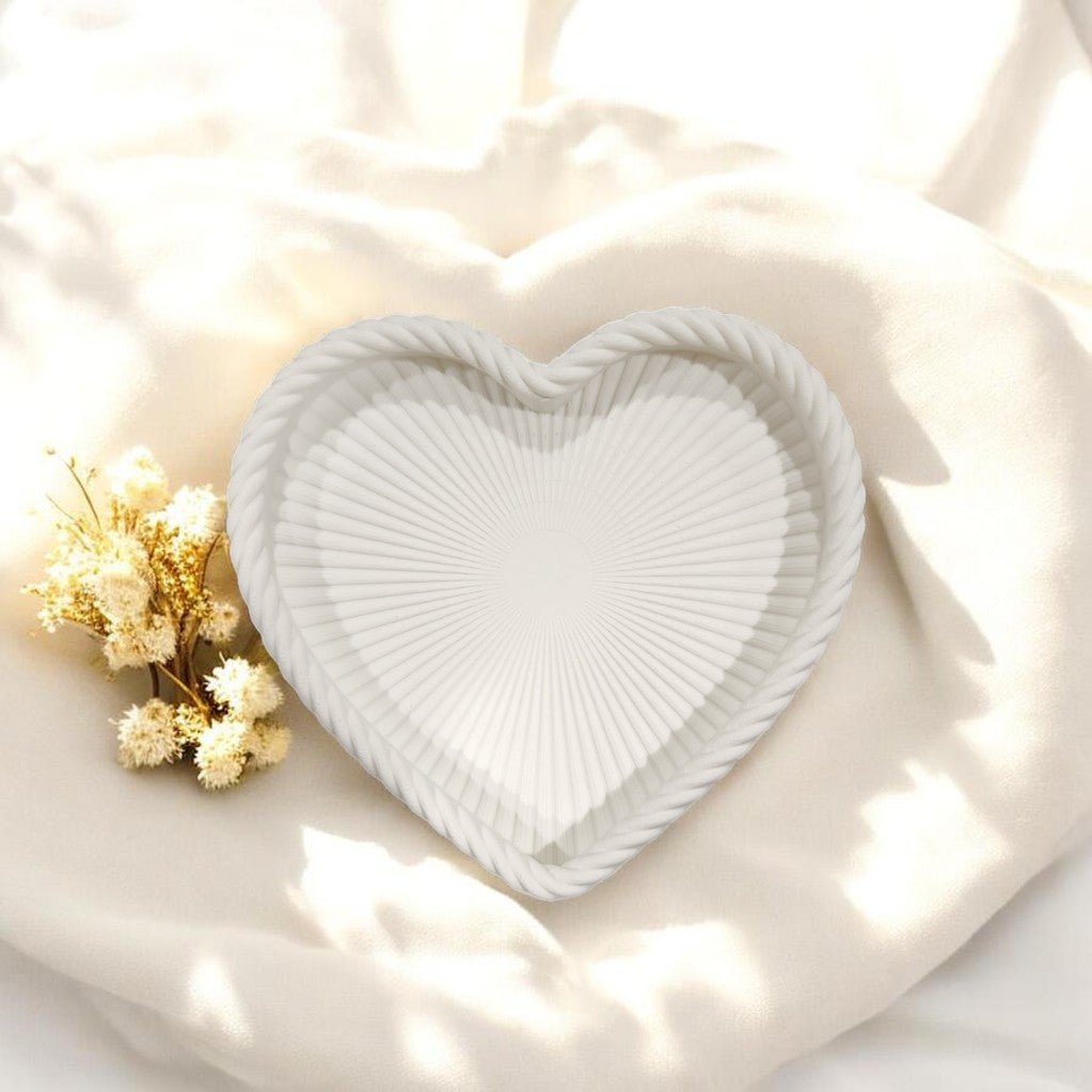 The Coastal Heart Tray in Matte White by Mirabilia Boutique, featuring a seashell texture, sits on soft cream fabric next to dried yellow flowers, illuminated by bright natural light.