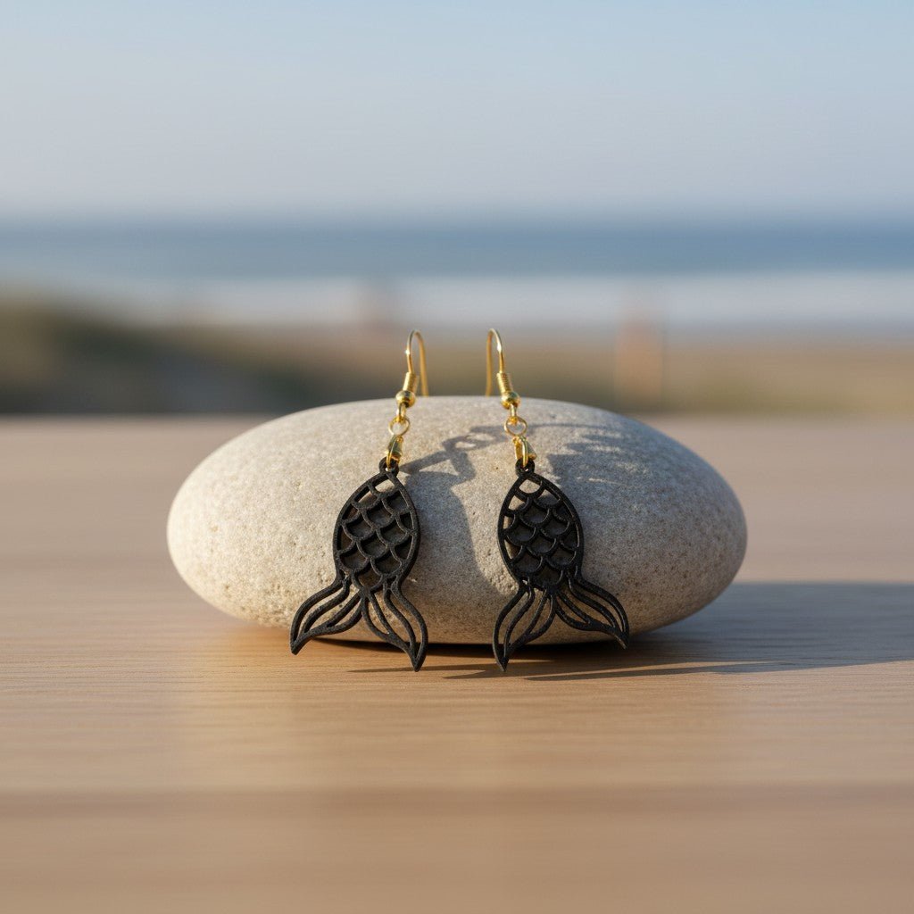 A pair of Mermaid Tail Earrings | Lasercut Walnut Wood by Mirabilia Boutique hang from a smooth stone on wood, with the beach and ocean blurred in the background—ideal coastal jewelry for ocean lovers.