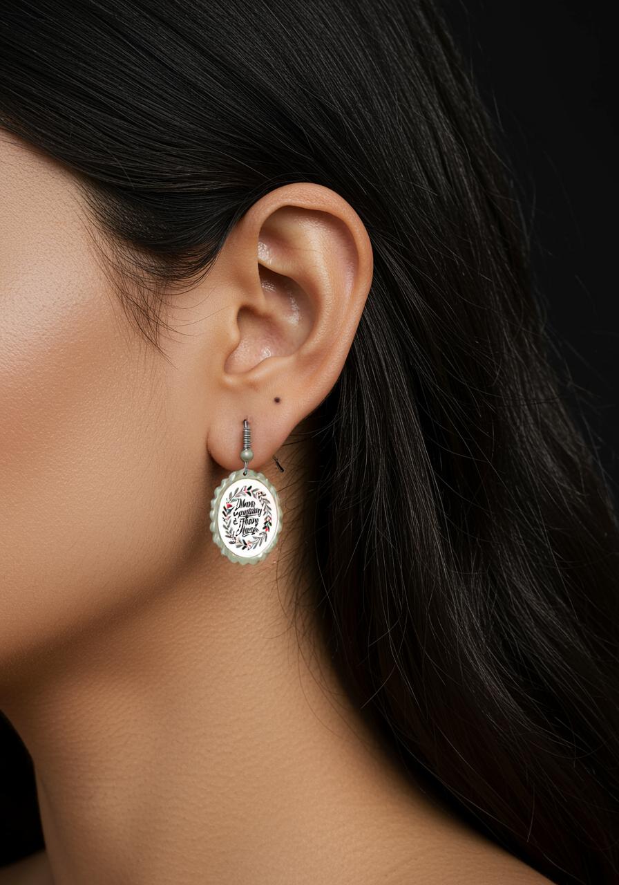 A close-up of a woman's ear showcases Mirabilia Boutique's Merry Everything Earrings: a larger laurel wreath earring with calligraphy and a silver stud. Her dark hair is tucked back, highlighting this festive holiday gift for her.
