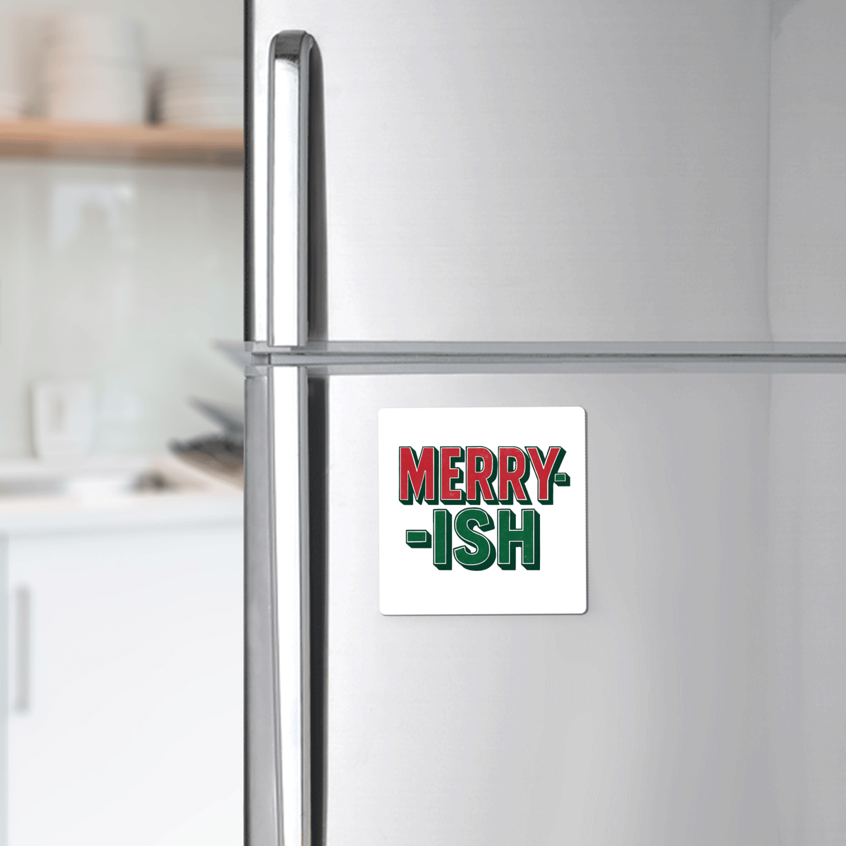 A white refrigerator features the Merry-ish Magnet by Mirabilia Boutique, a funny holiday fridge decor with bold red and green lettering; a blurred kitchen appears in the background.