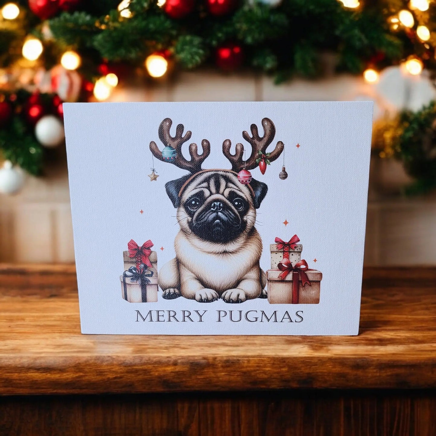 A Merry Pugmas 10x8 Canvas Sign by Mirabilia Boutique is displayed on a wooden table. The canvas features an illustration of a pug wearing antlers and surrounded by wrapped gifts, with the text "Merry Pugmas" prominently displayed. In the background, framed holiday decor and a decorated Christmas tree are partially visible.