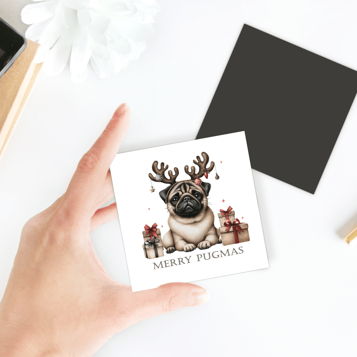 A hand displays the Merry Pugmas Magnet by Mirabilia Boutique, featuring a festive pug in reindeer antlers surrounded by gifts—perfect as a cute pug Christmas gift or cheerful holiday decor.