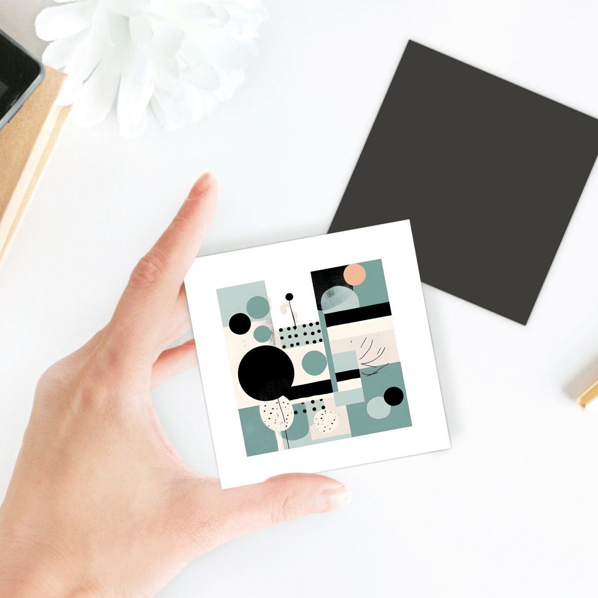 A hand holds the Harbor Morning Mid Century Modern Magnet by Mirabilia Boutique, featuring abstract geometric art in muted hues—ideal as geometric decor; beside it, a dark square magnet and a white flower rest on the white surface.