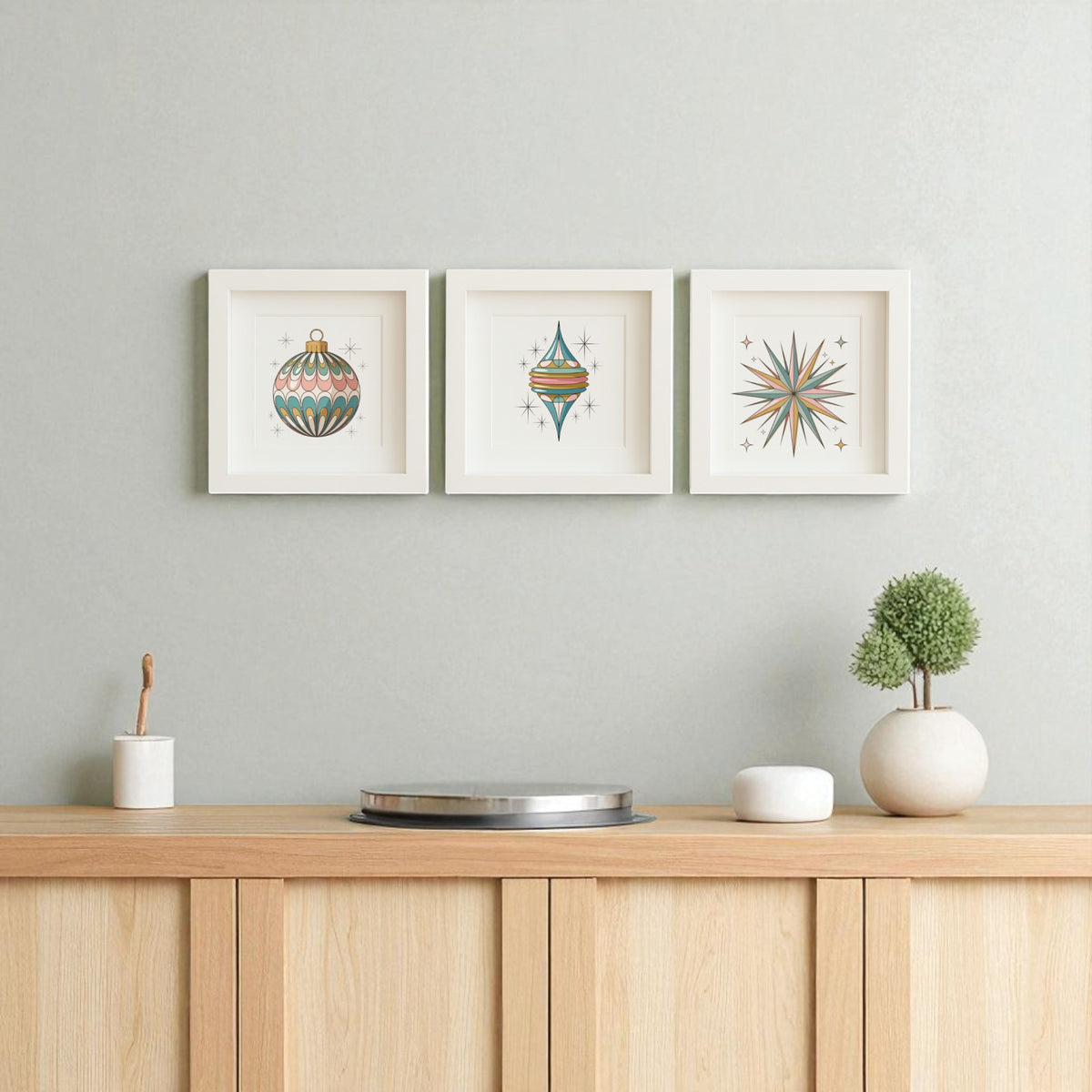 A minimalist wooden sideboard displays a round tray, white pot, and plant. Above, three Mirabilia Boutique "Mid-Century Round Ornament Art | Retro Holiday Shadowbox" prints add retro holiday charm to the light gray wall.