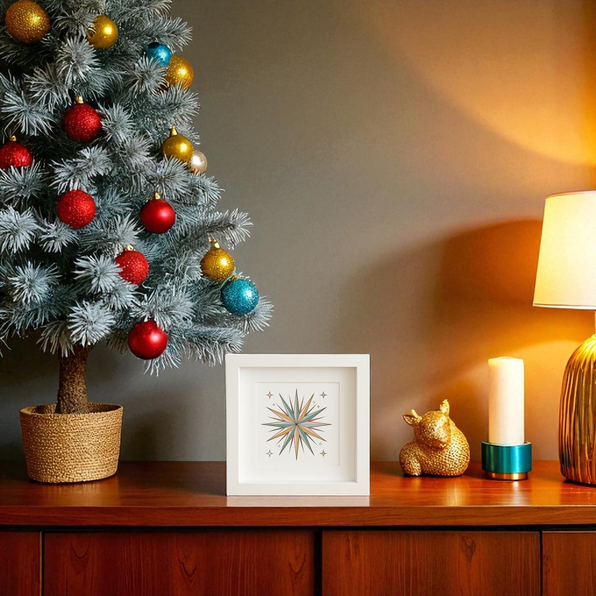 A small decorated Christmas tree stands on a wooden sideboard next to Mirabilia Boutique's Atomic Starburst Ornament Art (Retro Holiday Shadowbox), a gold deer, a white candle, and a lit table lamp against a warm neutral wall.