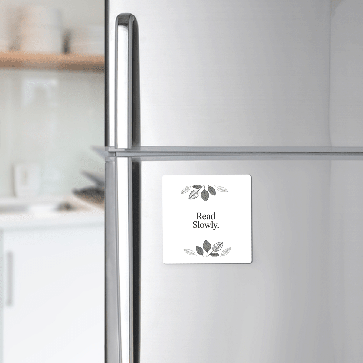 The Read Slowly Book Lover Magnet by Mirabilia Boutique—a white square magnet with green leaves and the words "Read Slowly."—adds literary charm to any silver refrigerator. White dishes are softly blurred in the background.