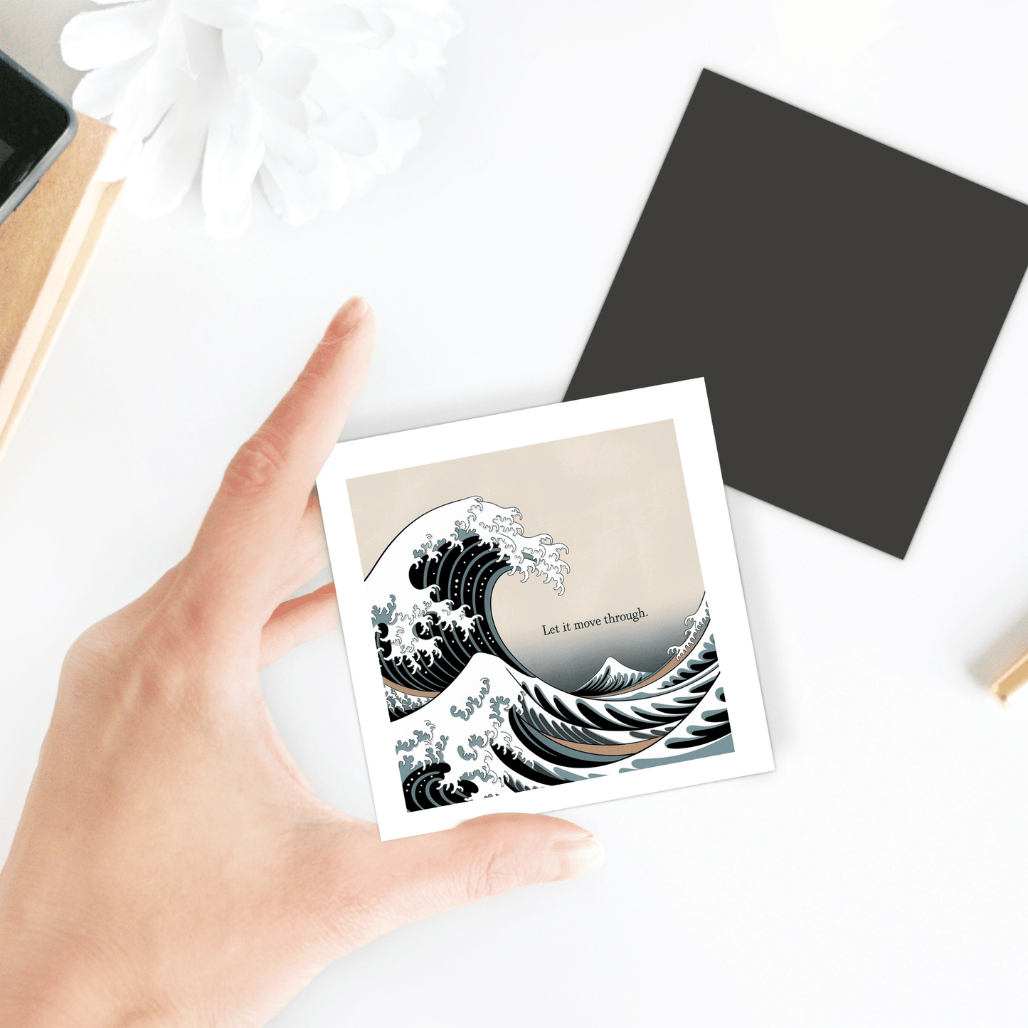 A hand holds the "Let It Move Through Magnet – Storm & Stillness Quote" by Mirabilia Boutique, featuring a wave illustration. The magnet rests on a white surface with a flower, a black square, and other nearby items.