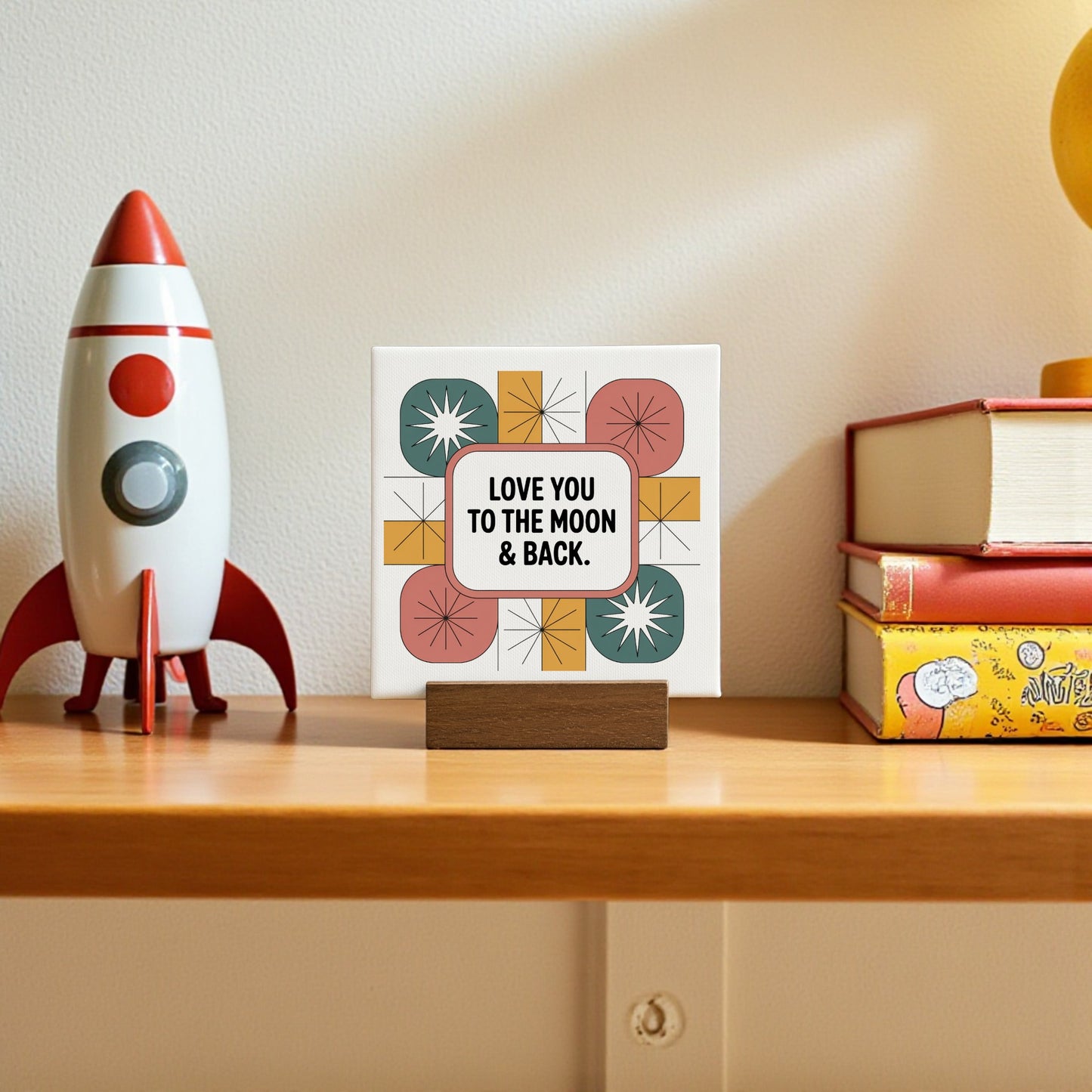 The Mirabilia Boutique "Love You to the Moon & Back" 4x4 mini canvas retro sign on a shelf with a toy rocket, stacked books, and a yellow mug makes a charming home decor love gift.
