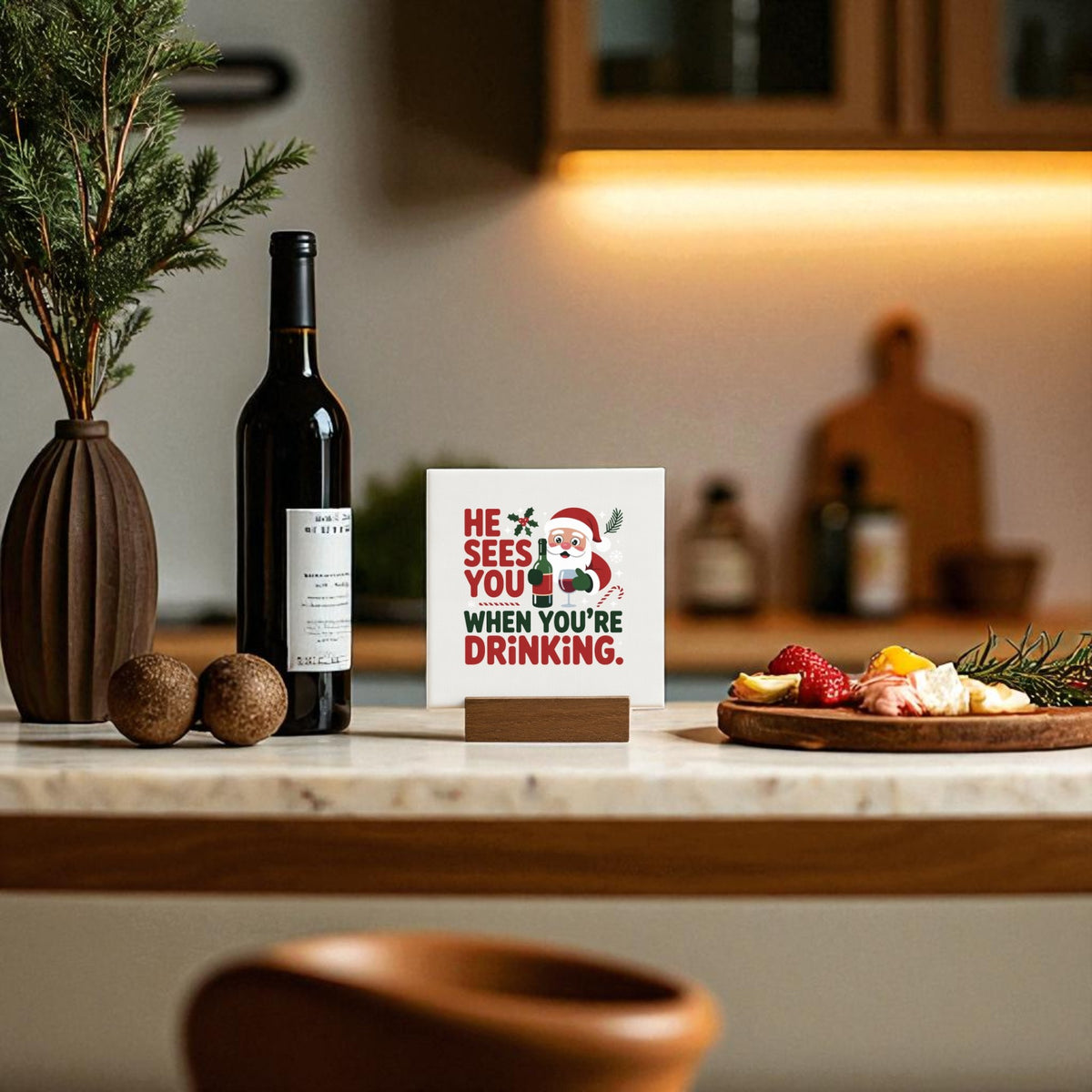 A kitchen counter features a wine bottle, a vase with greenery, fruit and cheese, and Mirabilia Boutique’s “He Sees You When You’re Drinking” funny mini Christmas sign—an ideal touch for your holiday bar cart décor.