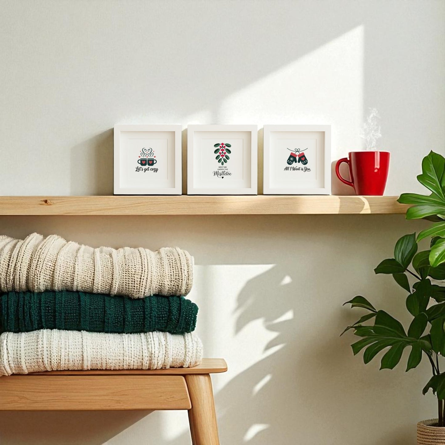 The Romantic Holiday Wall Art Trio | 5x5 Framed Gift Set by Mirabilia Boutique is displayed on a wooden shelf with a red mug and green plant, while cream and dark green knitted blankets rest below on a bench in sunlit shadows.