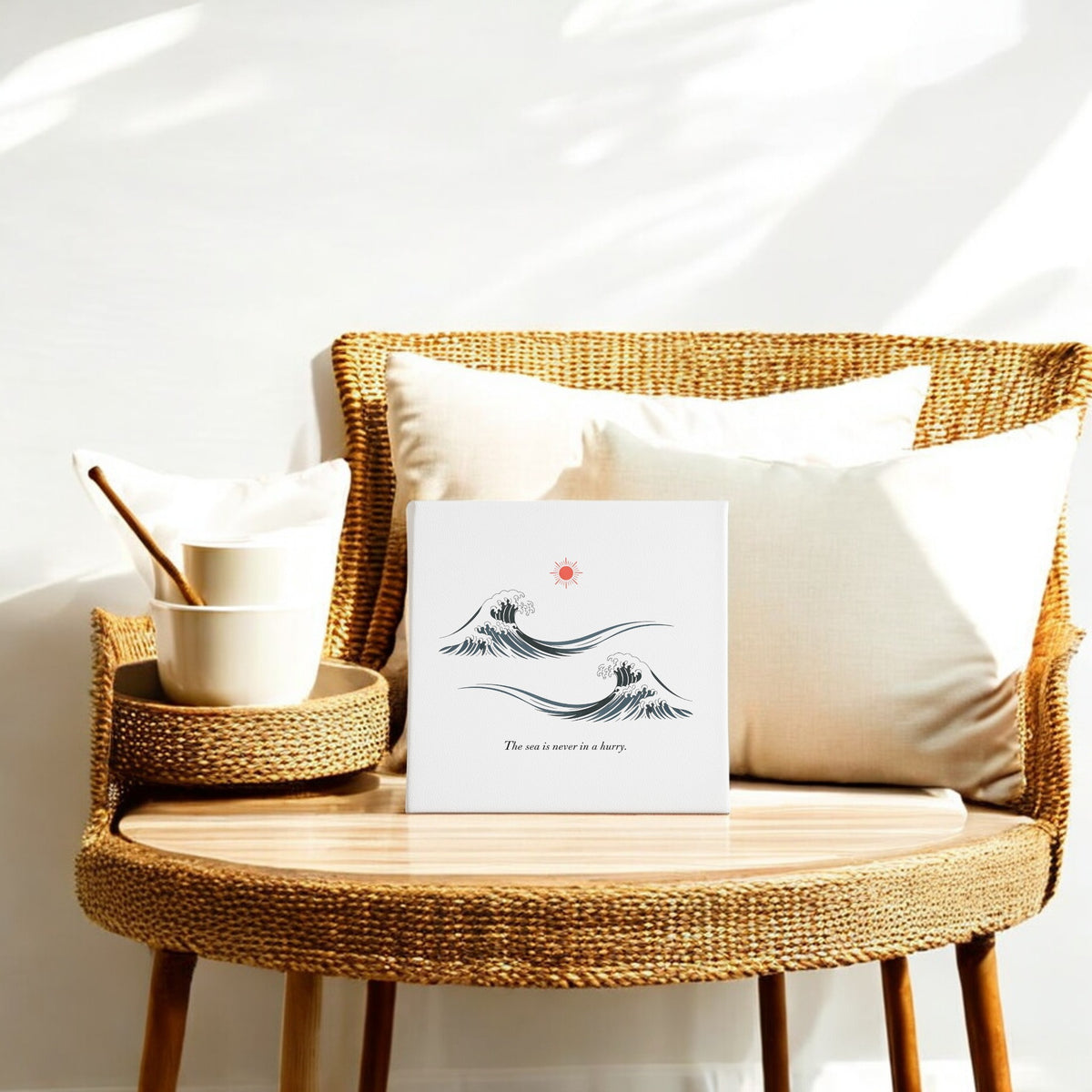 A rattan chair with white cushions sits beside a wooden table topped with stacked bowls and Mirabilia Boutique’s "The Sea Is Never in a Hurry Canvas – Serene Ocean Calm." Sunlight casts soft shadows on the wall behind.