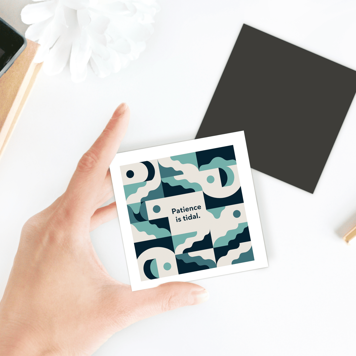 A hand holds the Mirabilia Boutique "Patience Is Tidal Magnet – Ocean-Inspired Calm Quote," featuring blue and cream wave art and the words "Patience is tidal." This inspirational fridge magnet adds a calming touch to any space.