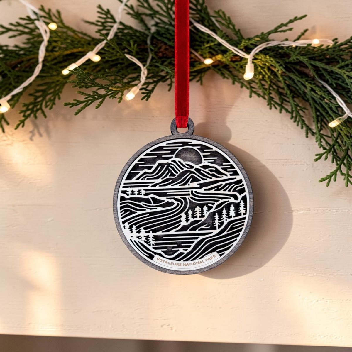 The Mirabilia Boutique Voyageurs National Park Line Art Christmas Ornament is an oval, handcrafted wooden piece featuring layered wood art with a black and white scenic design of trees and water, plus "Voyageurs National Park" text at the bottom. It hangs from a red ribbon, while pine branches with string lights are shown above.