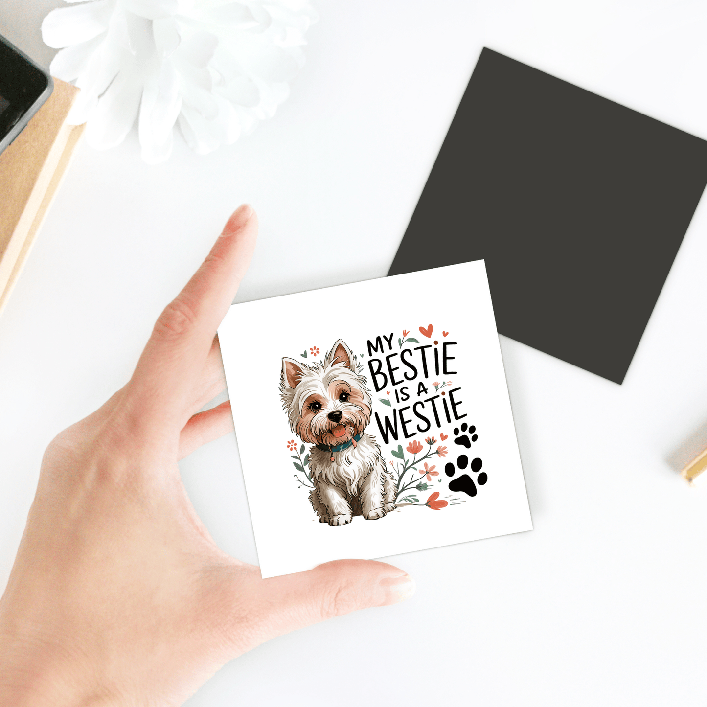 A hand displays the Westie Dog Magnet by Mirabilia Boutique, a square card with a cute Westie, florals, paw prints, and "My Bestie is a Westie." Ideal as a West Highland White Terrier gift or dog lover magnet. A white flower and black square are nearby.