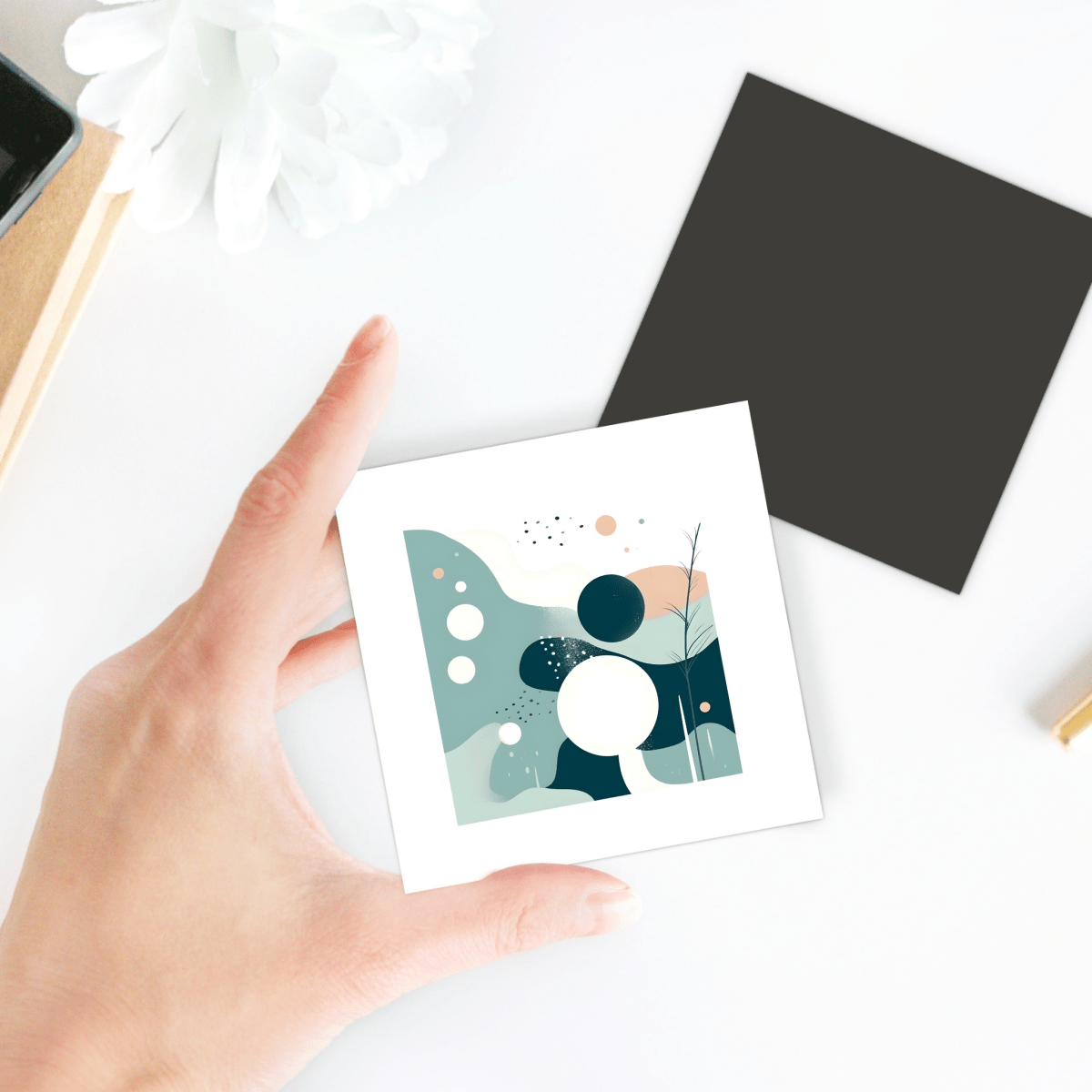 A hand holds the Quiet Tides Mid Century Modern Magnet by Mirabilia Boutique, featuring abstract teal, white, navy, and peach shapes. Nearby on the white desk are a black square card, flower, and pen.
