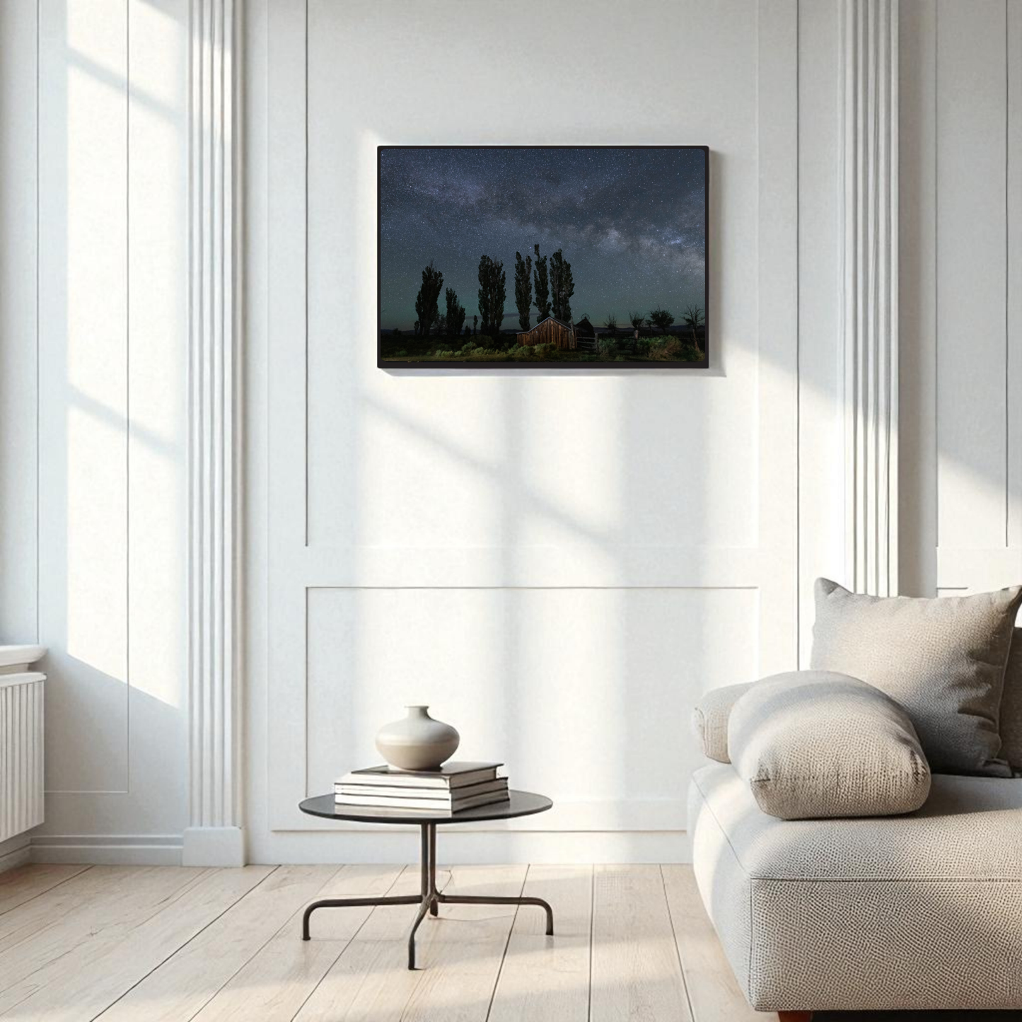 A modern living room with a beige sofa, round coffee table, and a vase. On the white paneled wall hangs the DeChambeau Ranch Milky Way Print | Framed Fine Art Print by Mirabilia Boutique—an archival-quality landscape photograph of trees beneath the Eastern Sierra night sky. Sunlight streams in from large windows.
