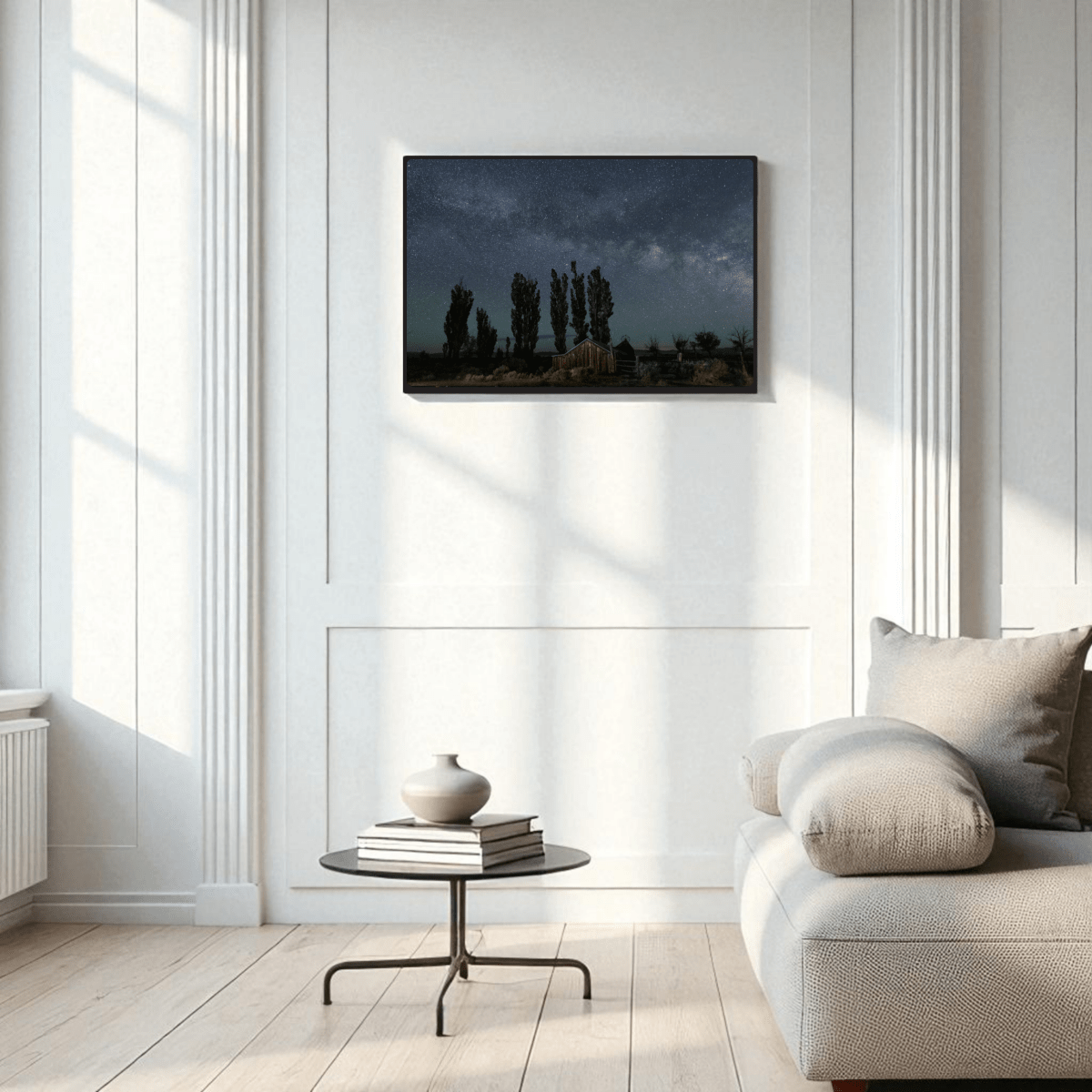 A modern living room with a beige sofa, round coffee table, and a vase. On the white paneled wall hangs the DeChambeau Ranch Milky Way Print | Framed Fine Art Print by Mirabilia Boutique—an archival-quality landscape photograph of trees beneath the Eastern Sierra night sky. Sunlight streams in from large windows.